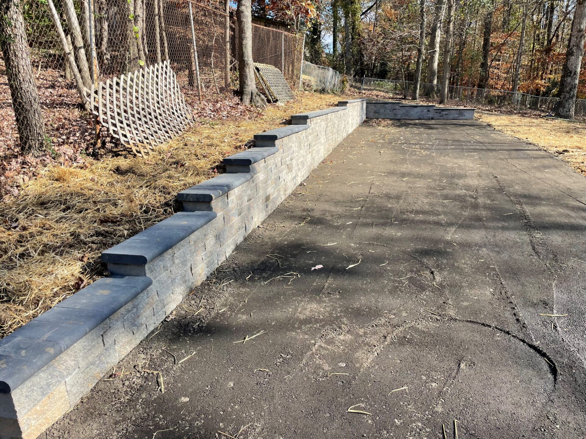 A paved pathway with a brick retaining wall and wooden fence panels, surrounded by trees and dried grass, under clear skies.
