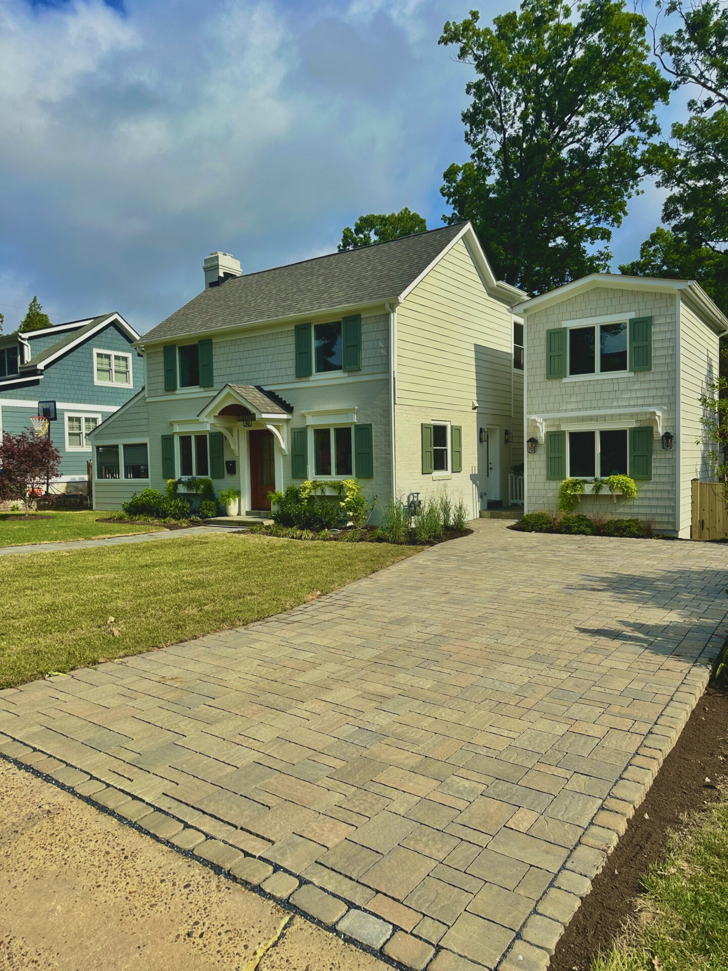 A suburban house with light siding, green shutters, large driveway, and lush lawn, set against a backdrop of trees and cloudy sky.