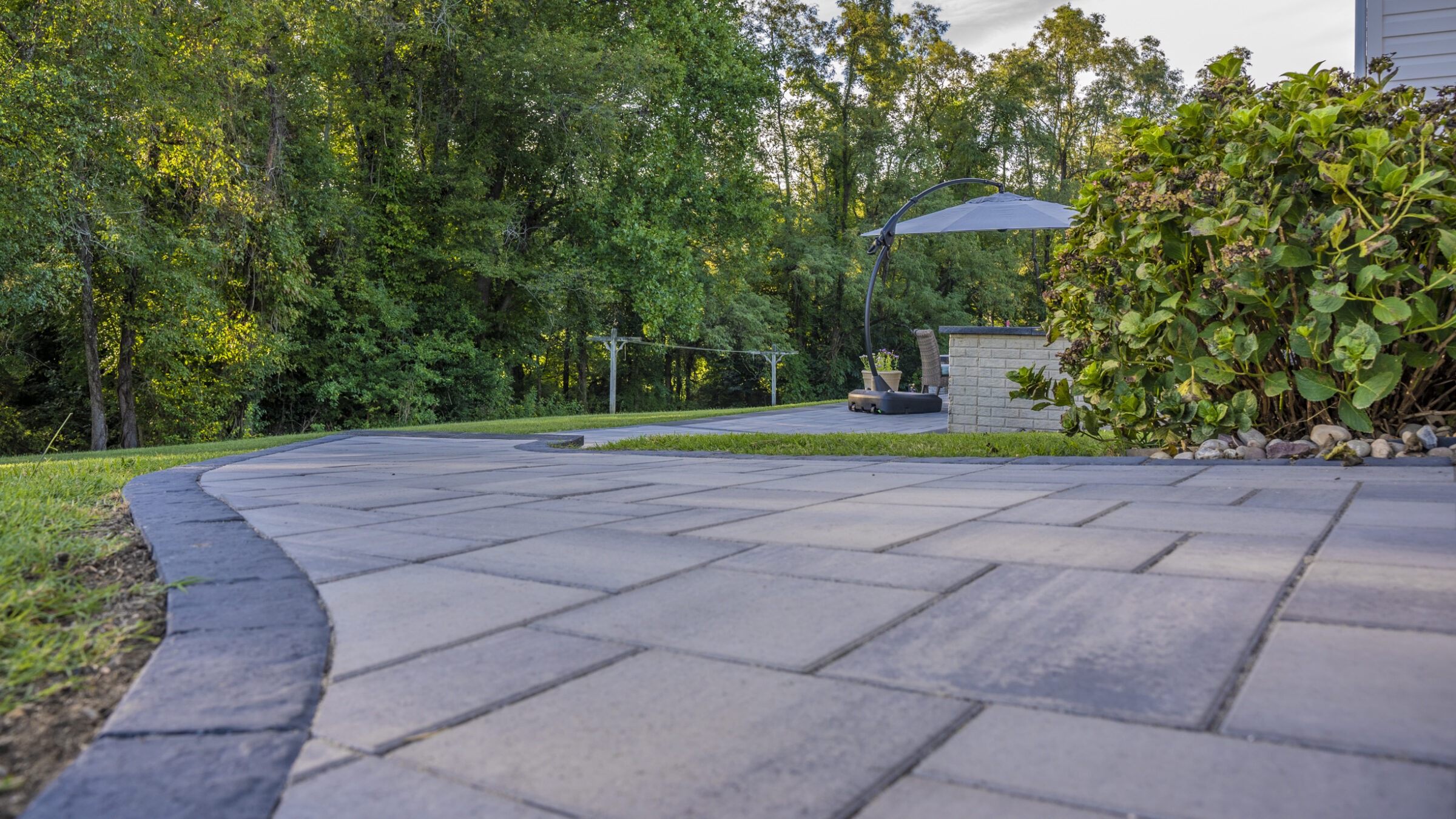 Curved stone pathway with adjacent greenery and trees, leading to a patio area featuring outdoor furniture and an umbrella under a clear sky.