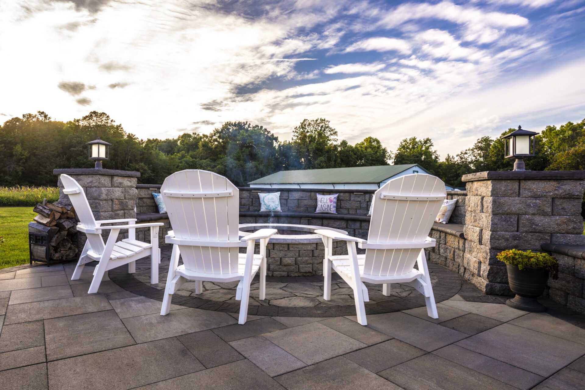 Three white chairs around a stone fire pit on a patio, surrounded by lush greenery and a partly cloudy sky.
