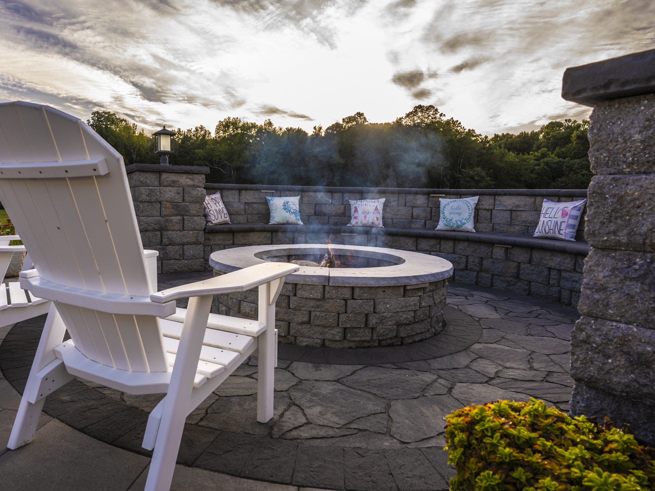 A serene patio features a cozy fire pit, white chairs, and colorful pillows against a lush, green backdrop under a dramatic sky.