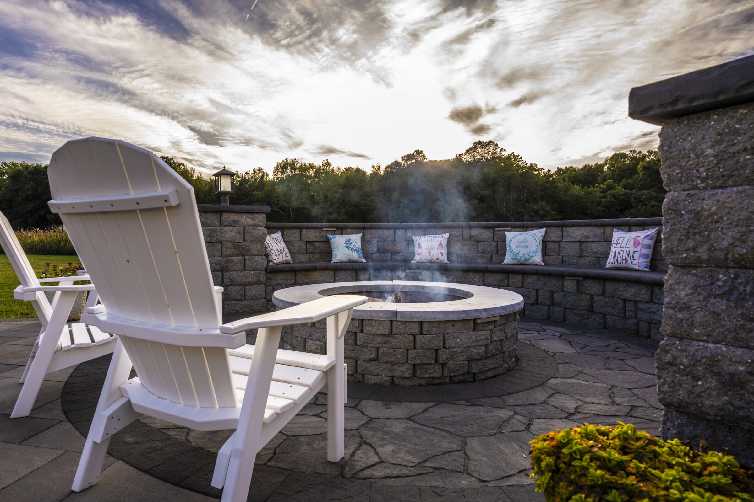 Patio scene with Adirondack chairs surrounding a stone fire pit. Cushions on a bench, cloudy sky, and trees in the background.