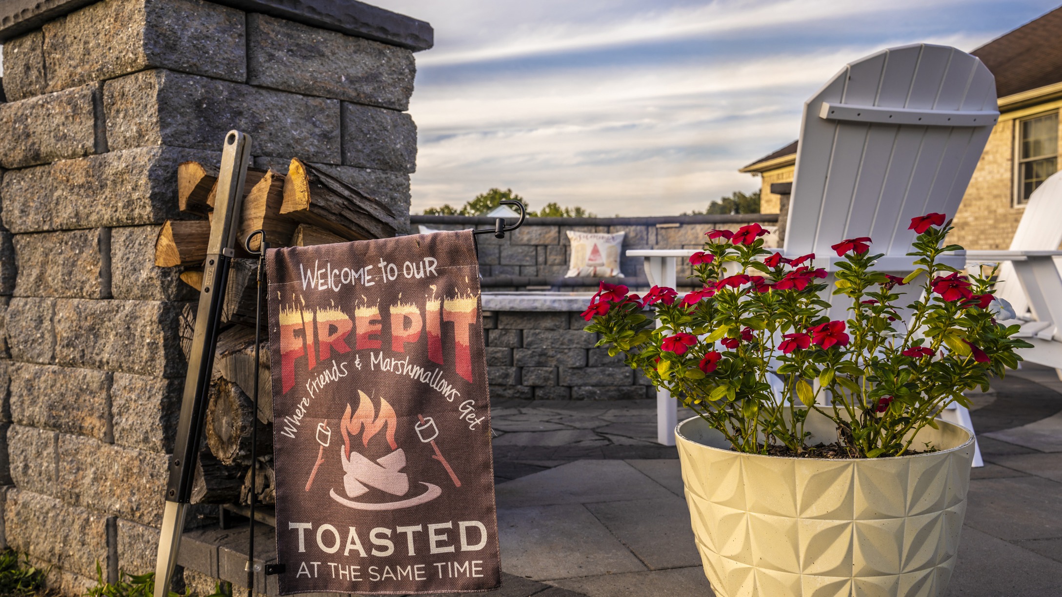 Stone patio with fire pit sign, white chairs, and a potted plant with red flowers, creating a cozy outdoor seating area.