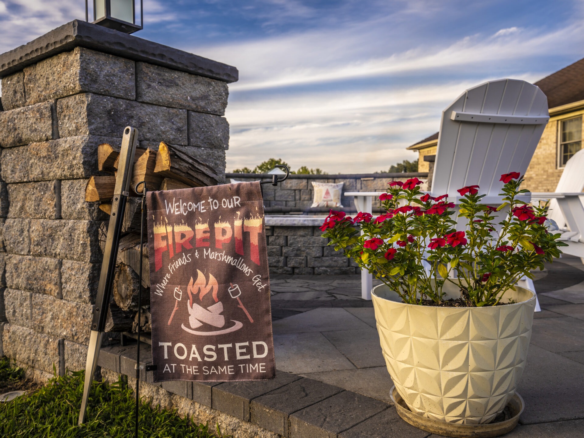Cozy outdoor fire pit area with red flowers, decorative sign, white chairs, and stacked firewood against a blue sky backdrop.