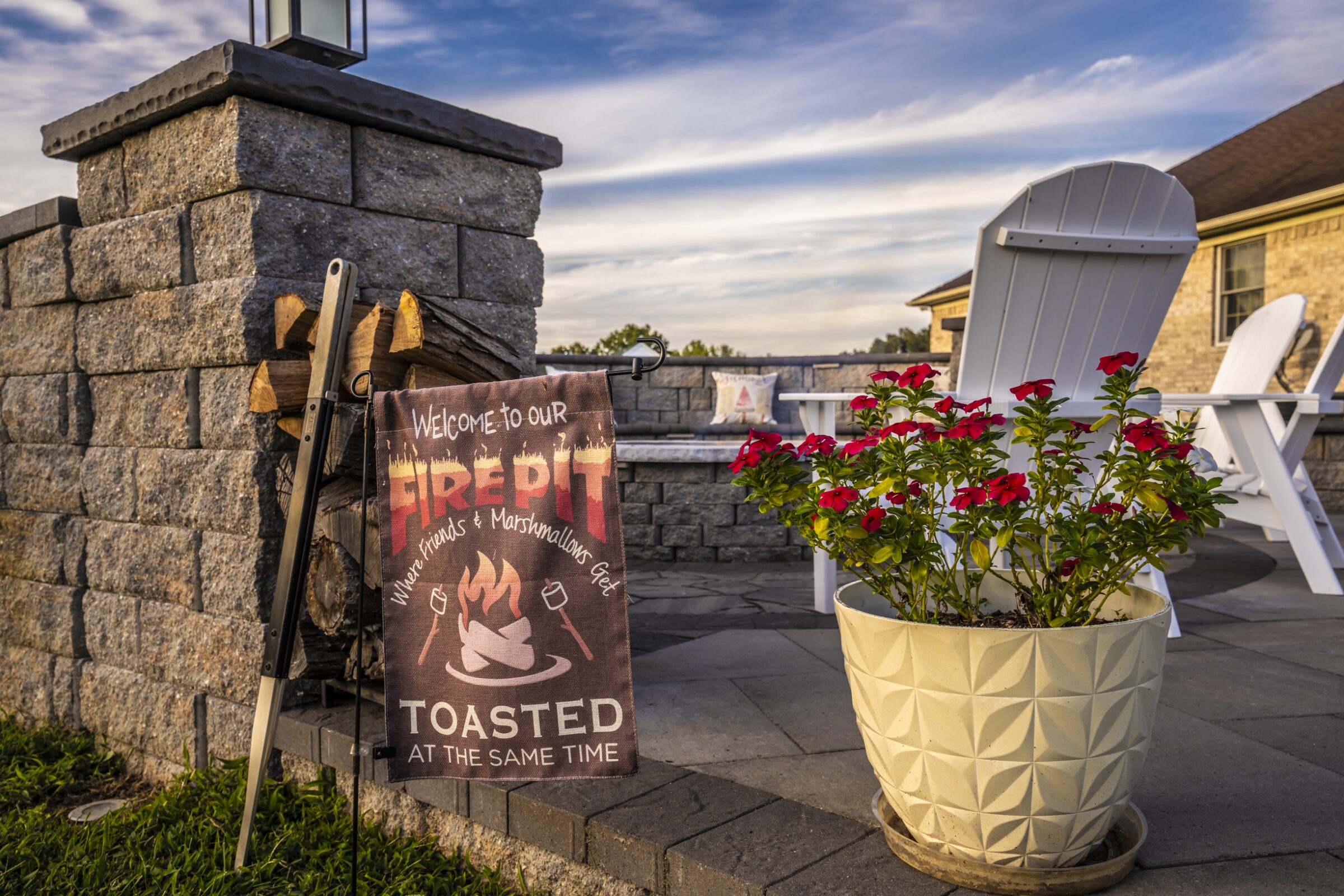 Outdoor fire pit area with a welcoming sign, white chairs, stacked wood, and a potted plant, set against a partly cloudy sky.