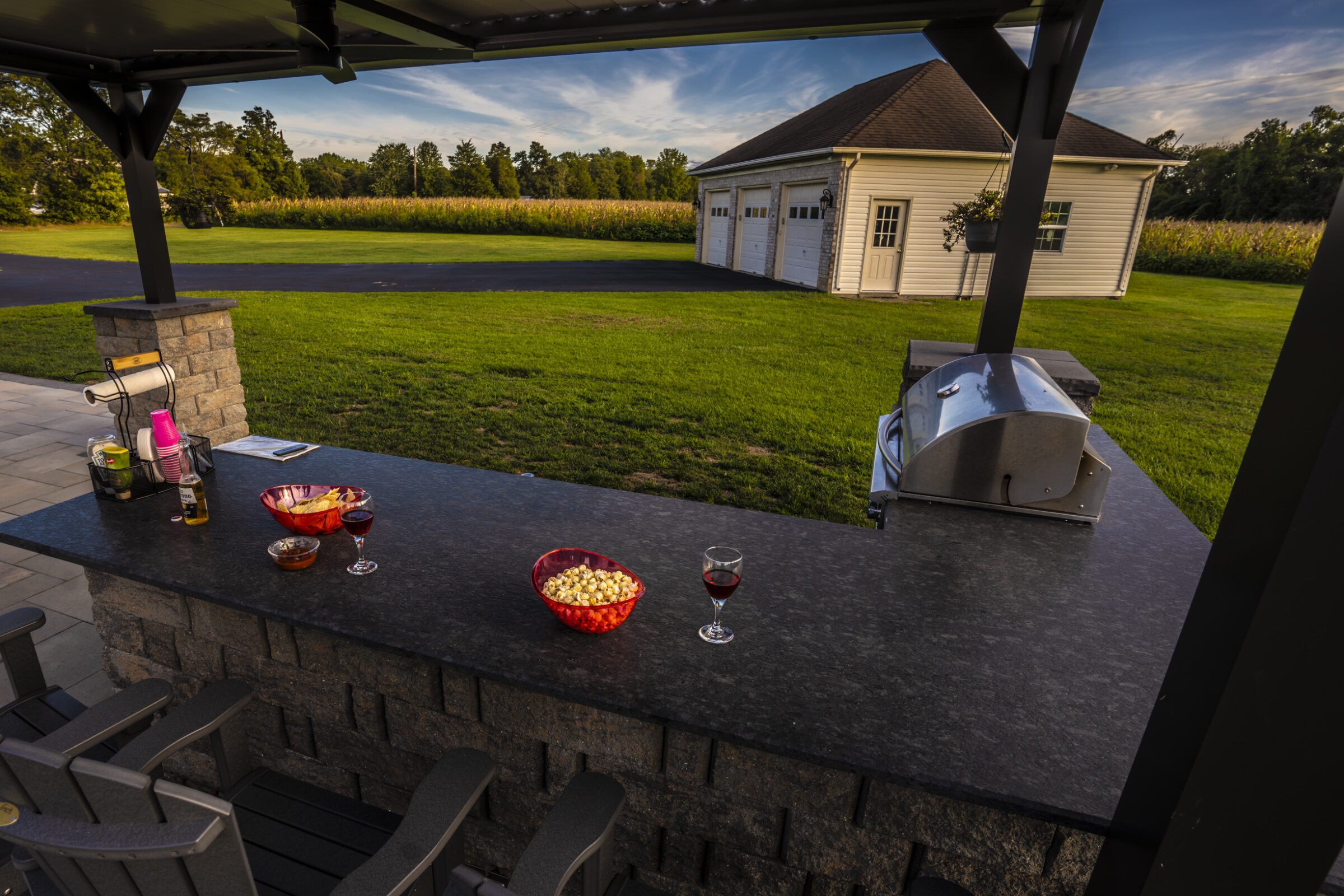 Outdoor patio with a grill, snacks, and drinks. A garage and green grass are visible under a clear sky.