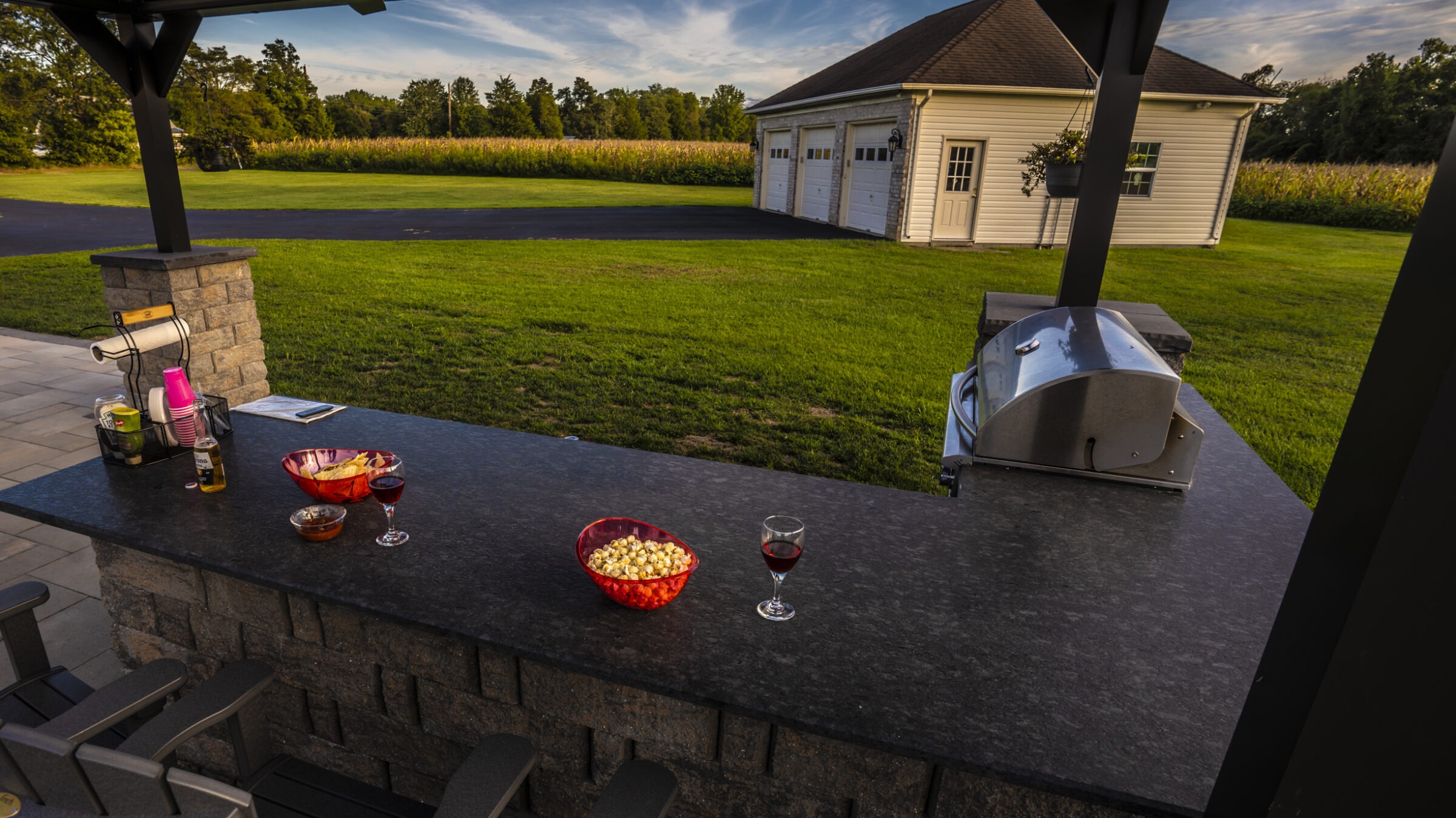 Outdoor kitchen with a countertop featuring snacks, drinks, and a grill. Lawn and garage in the background under a blue sky.