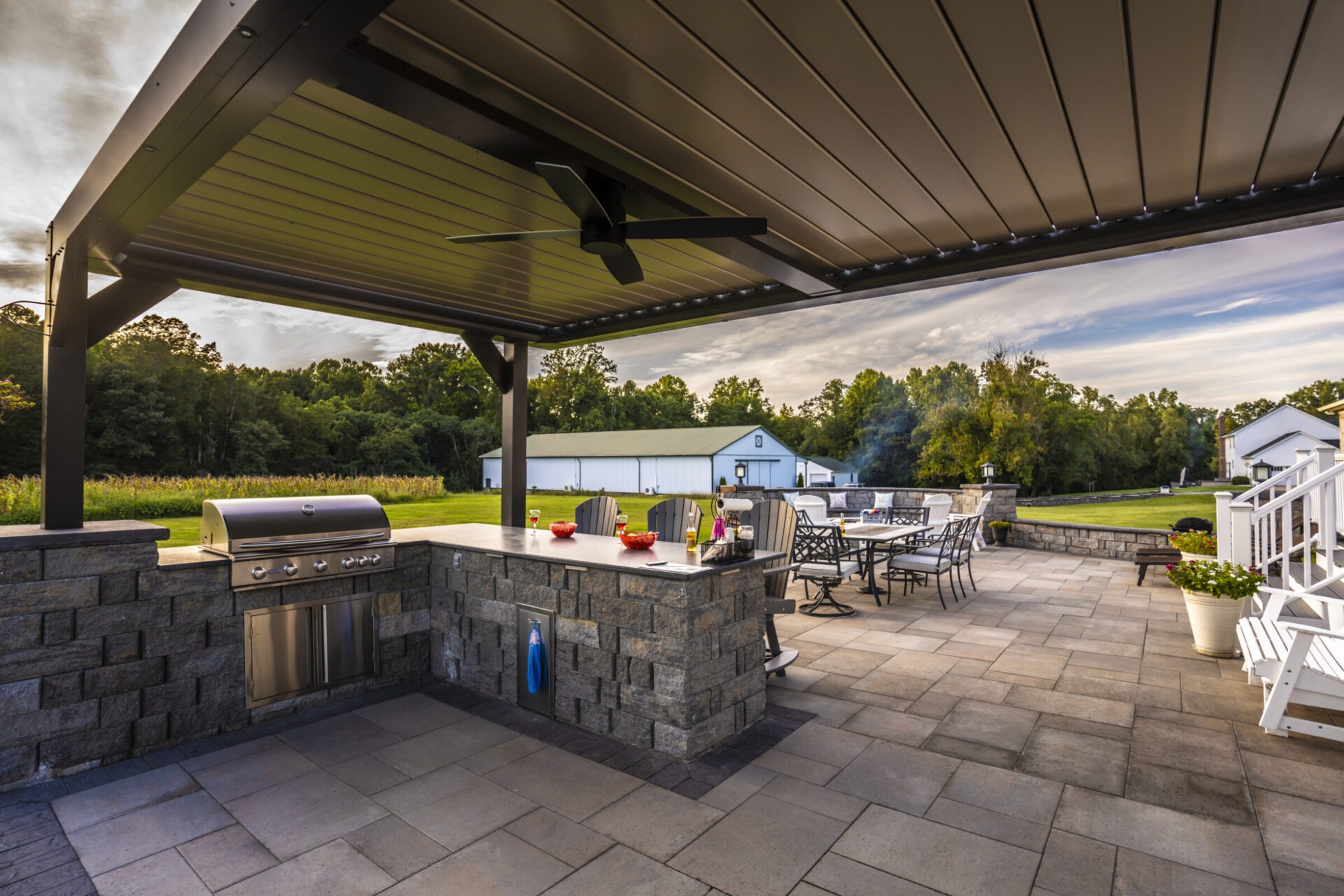 Outdoor patio with grilling area and dining furniture under a modern pergola. Surrounded by green lawn and trees, creating a peaceful setting.