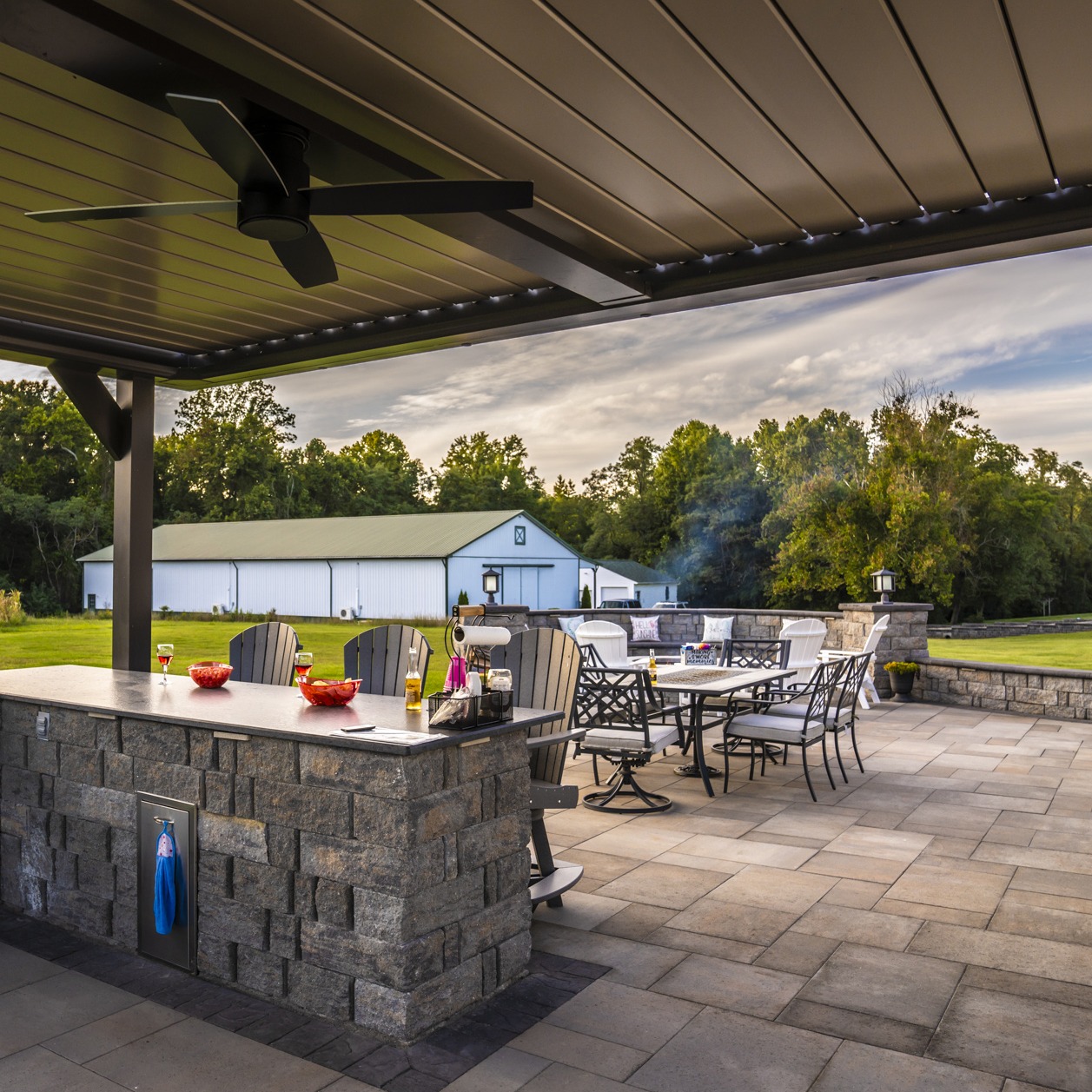 Outdoor patio with dining furniture and a bar area, under a pergola. Large white barn in background, surrounded by lush trees.