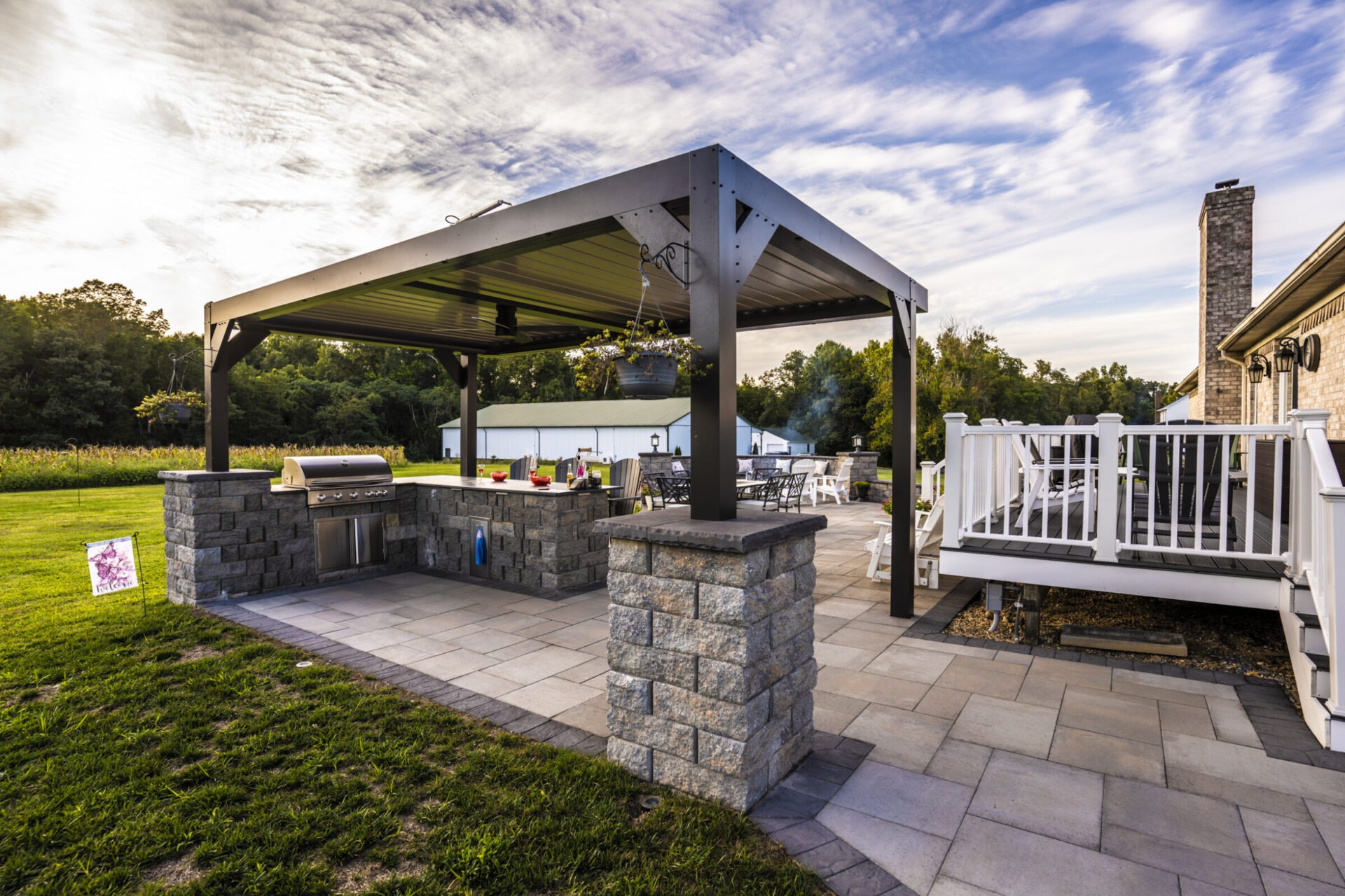 Outdoor patio with a pergola, grill, and seating area on a sunny day. Lush greenery and blue sky in the background.
