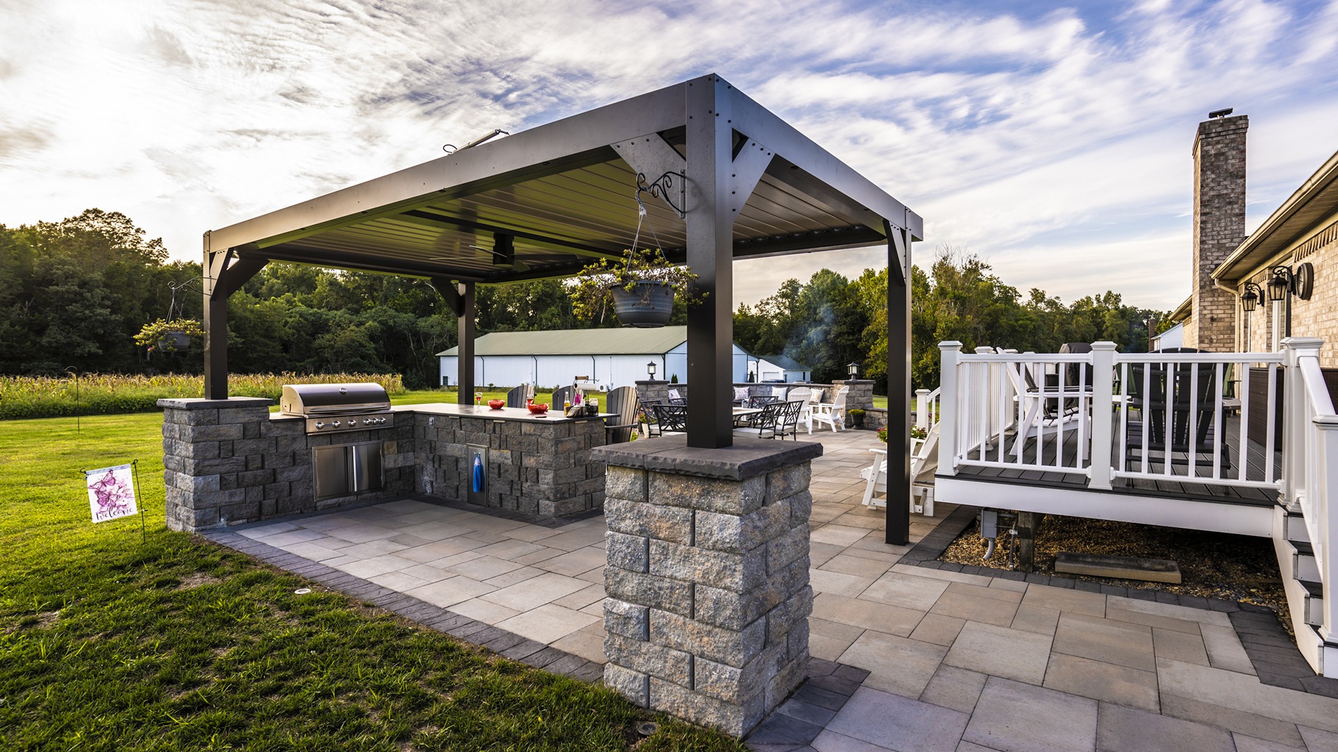 A spacious, modern outdoor patio with a pergola-covered grilling area and seating, surrounded by lush greenery under a partly cloudy sky.