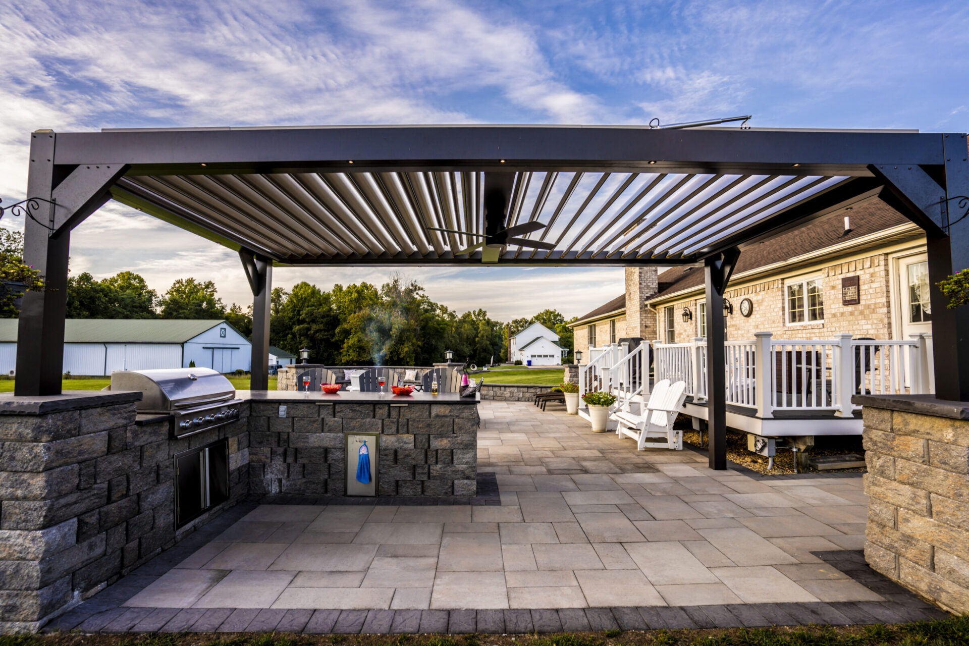 A backyard patio with a pergola, outdoor kitchen, and seating area. Modern furniture and barbecue grill under a blue sky.