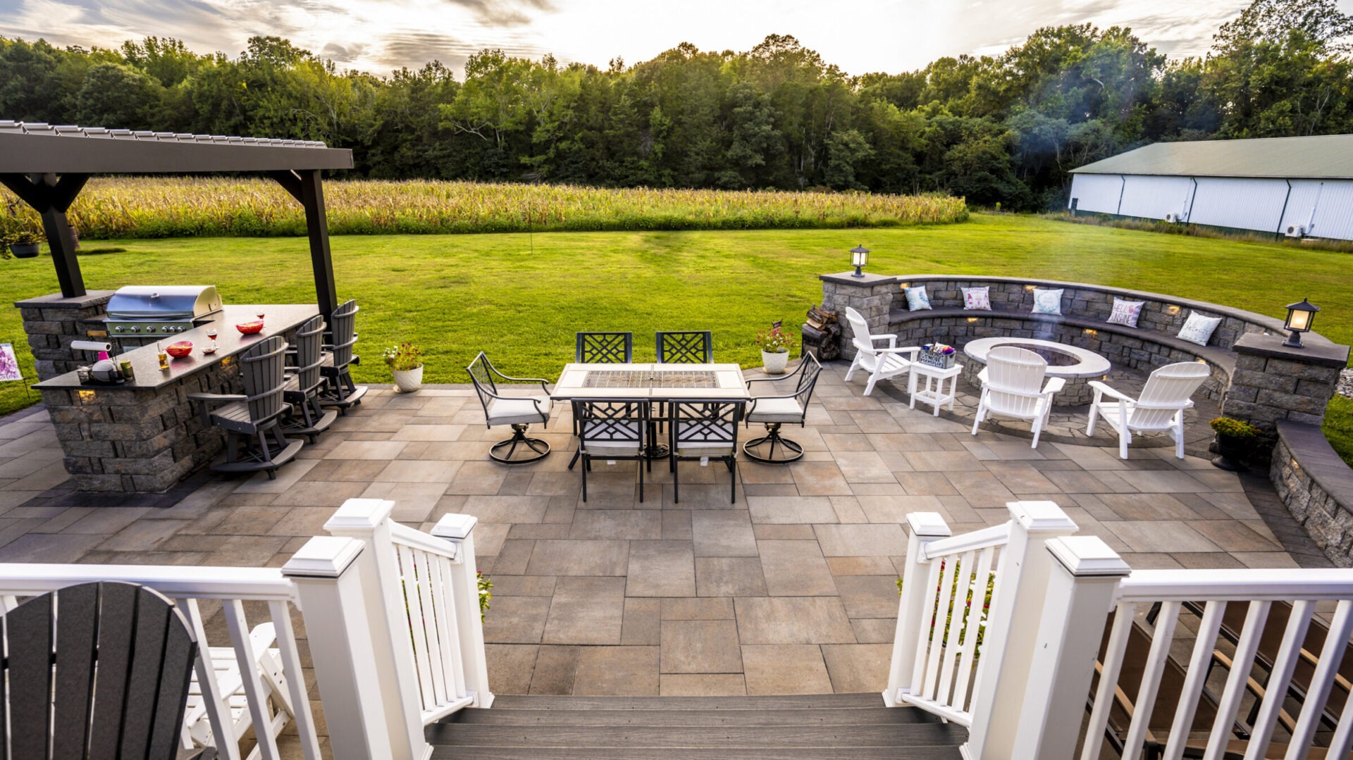 Spacious backyard patio with modern furniture, grill, and fire pit, overlooking a lush green lawn and forested area under a cloudy sky.
