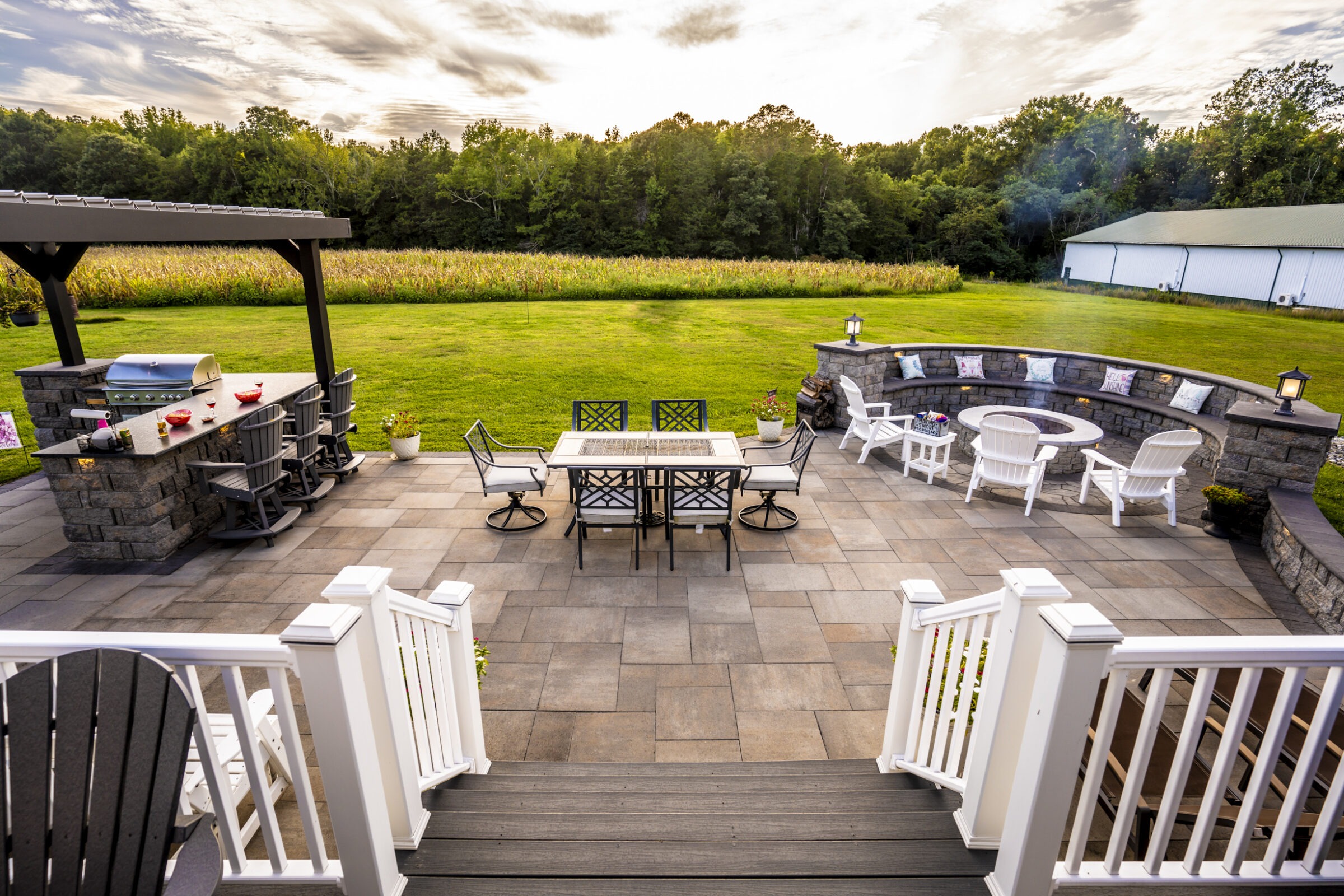Spacious backyard patio with a dining table, barbecue area, and fire pit surrounded by chairs. Overlooks a lush lawn and cornfield.