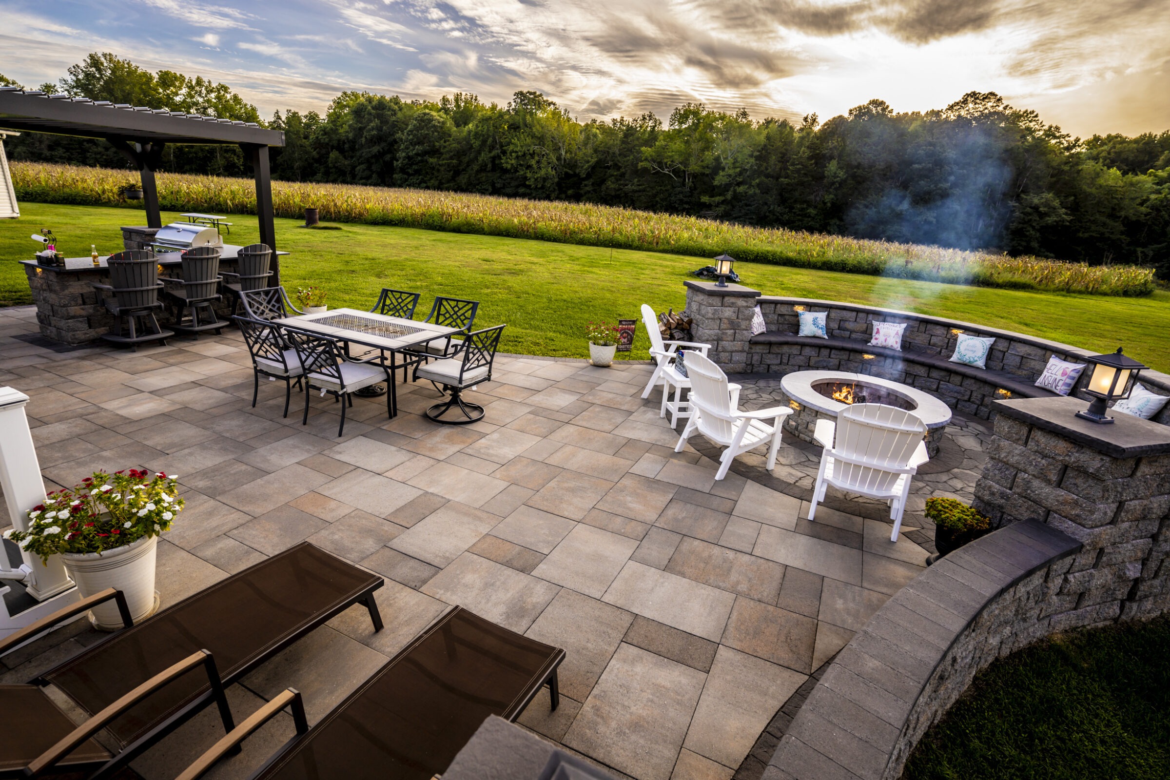 A scenic patio with a fire pit, dining area, and bar overlooks a lush green field and forest under a cloudy sky.