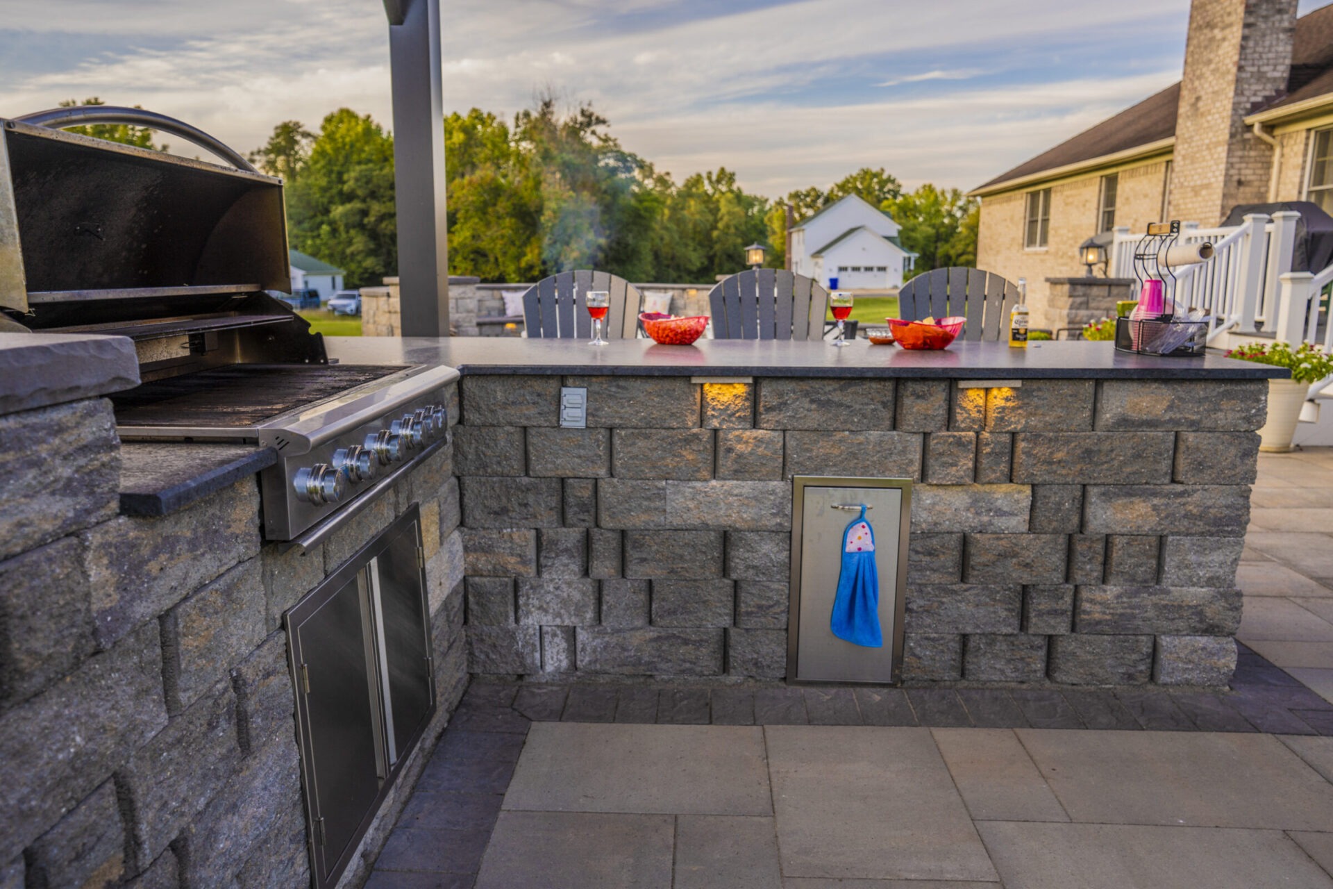 Outdoor kitchen with a stone counter, grill, drinks, and snacks. Background shows chairs, lush greenery, and a two-story house.