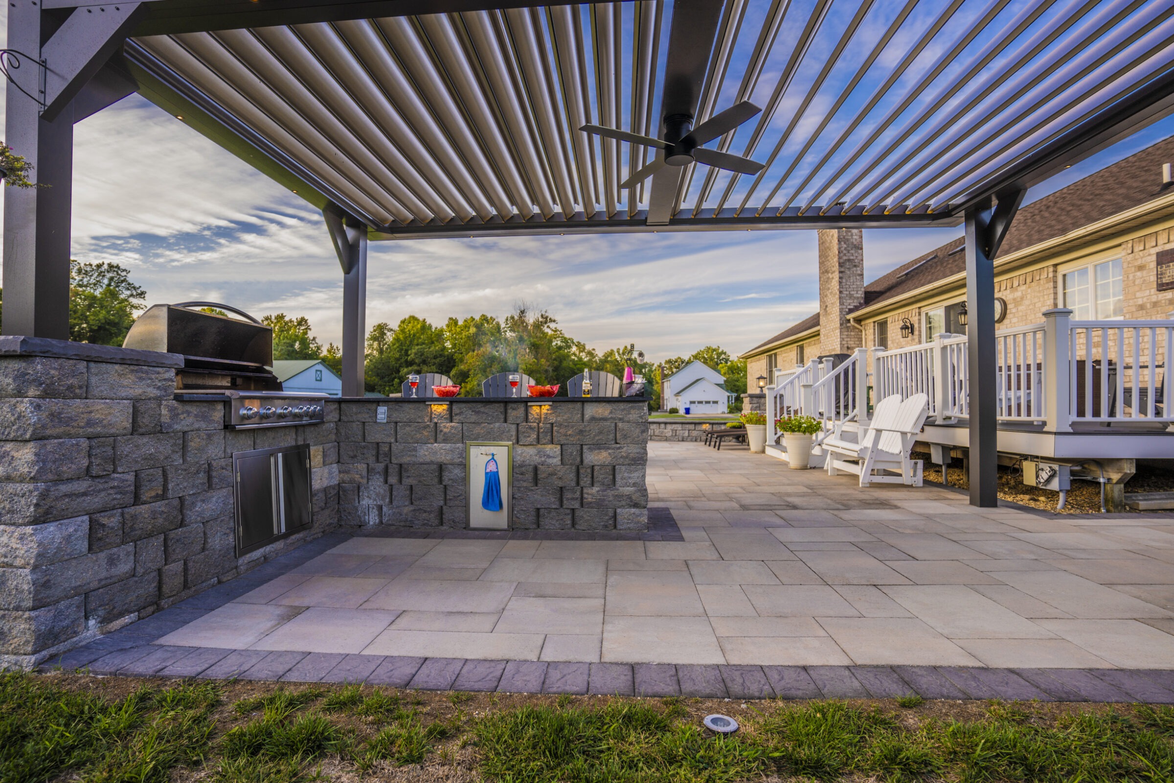 A modern outdoor patio features a stone grill, pergola, and seating area, set against a backdrop of greenery and suburban homes.