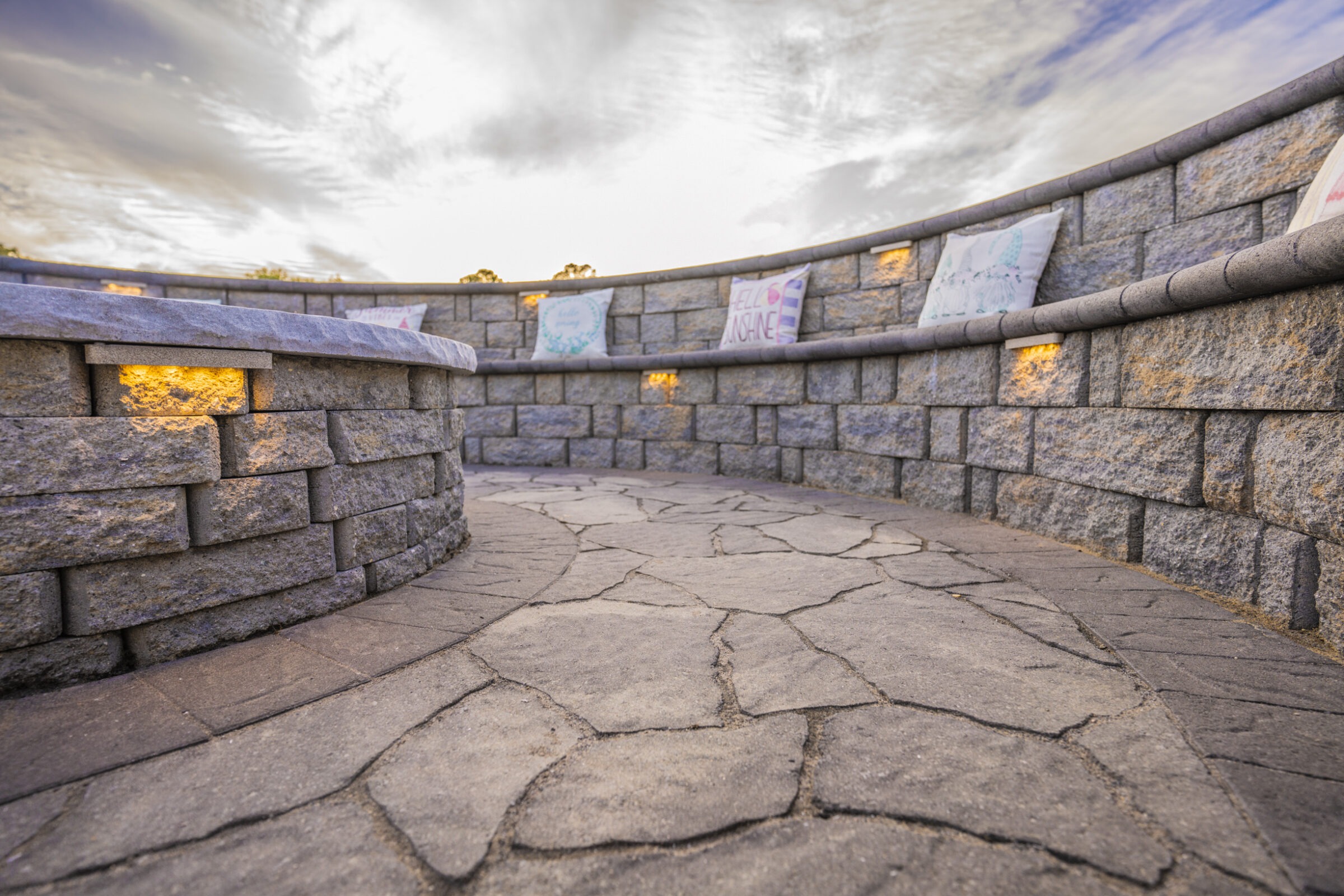 Circular stone bench with glowing lights, surrounded by stone walls. Cushions adorn the seats. Cloudy sky overhead creates a peaceful atmosphere.