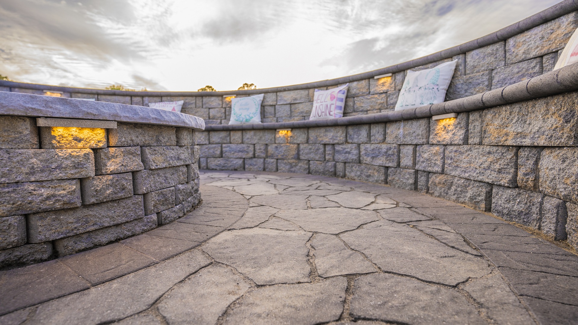 Circular stone seating area with decorative pillows under a cloudy sky. Warm lighting illuminates the walls, creating an inviting ambiance.