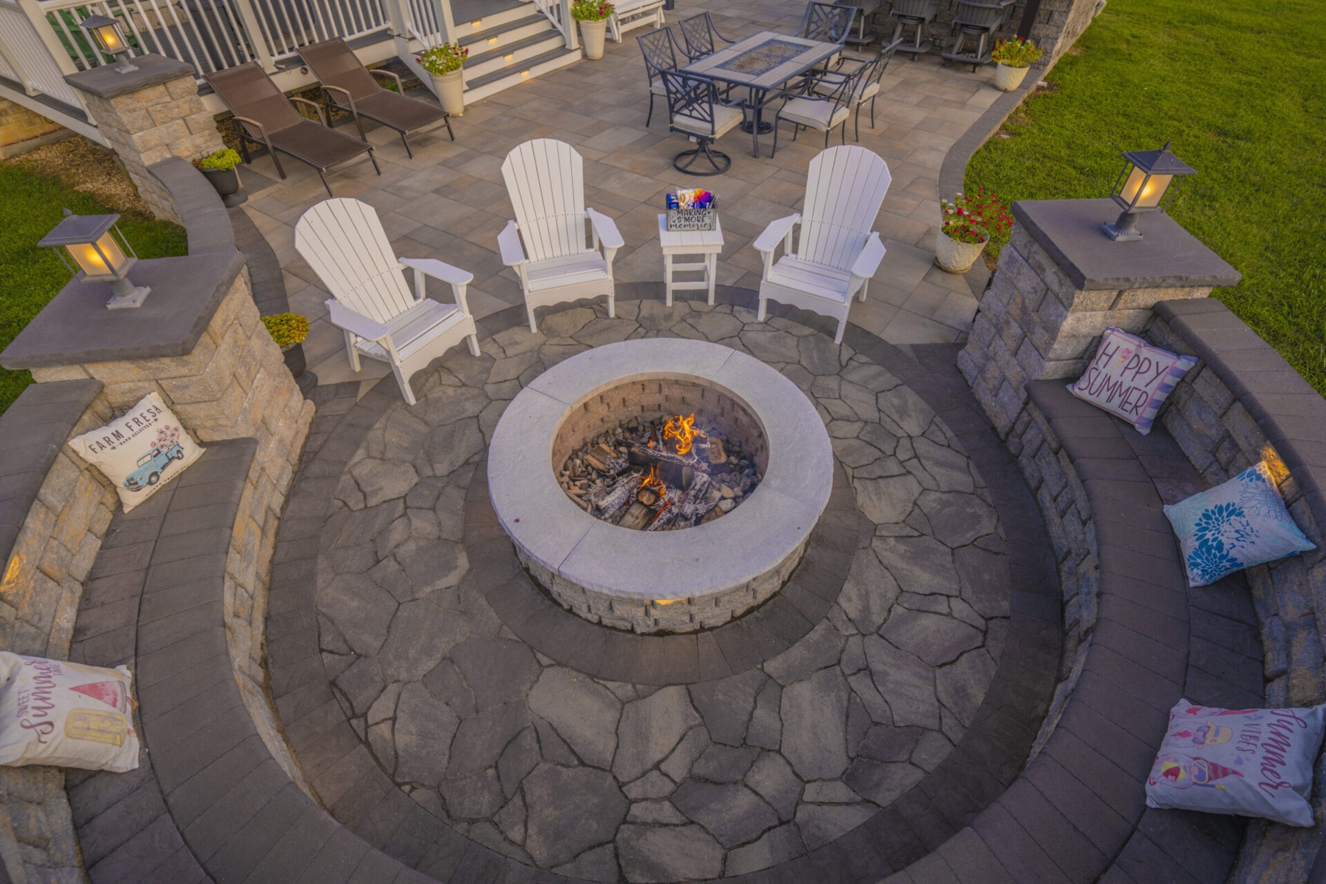 A cozy outdoor patio setup with a central fire pit, surrounded by chairs and decorative pillows, under warm lighting.
