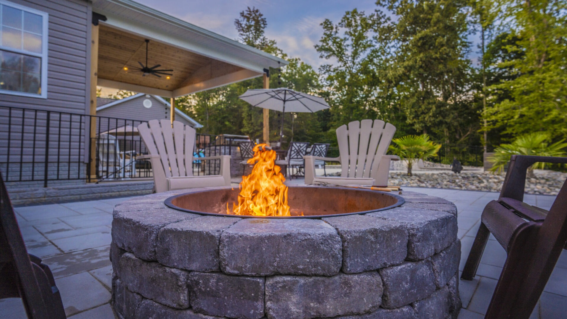 A cozy outdoor patio with a stone fire pit surrounded by chairs, adjacent to a covered porch and trees in the background.