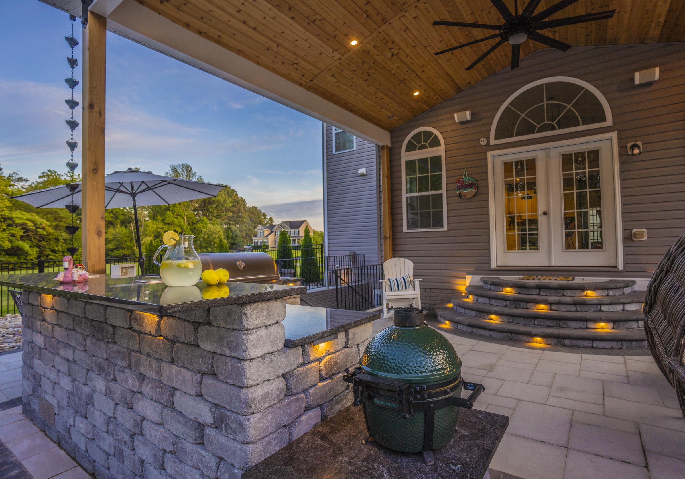 Covered patio with stone countertops, a grill, and outdoor seating. Lemonade pitcher on table. Cozy ambiance with evening lighting.