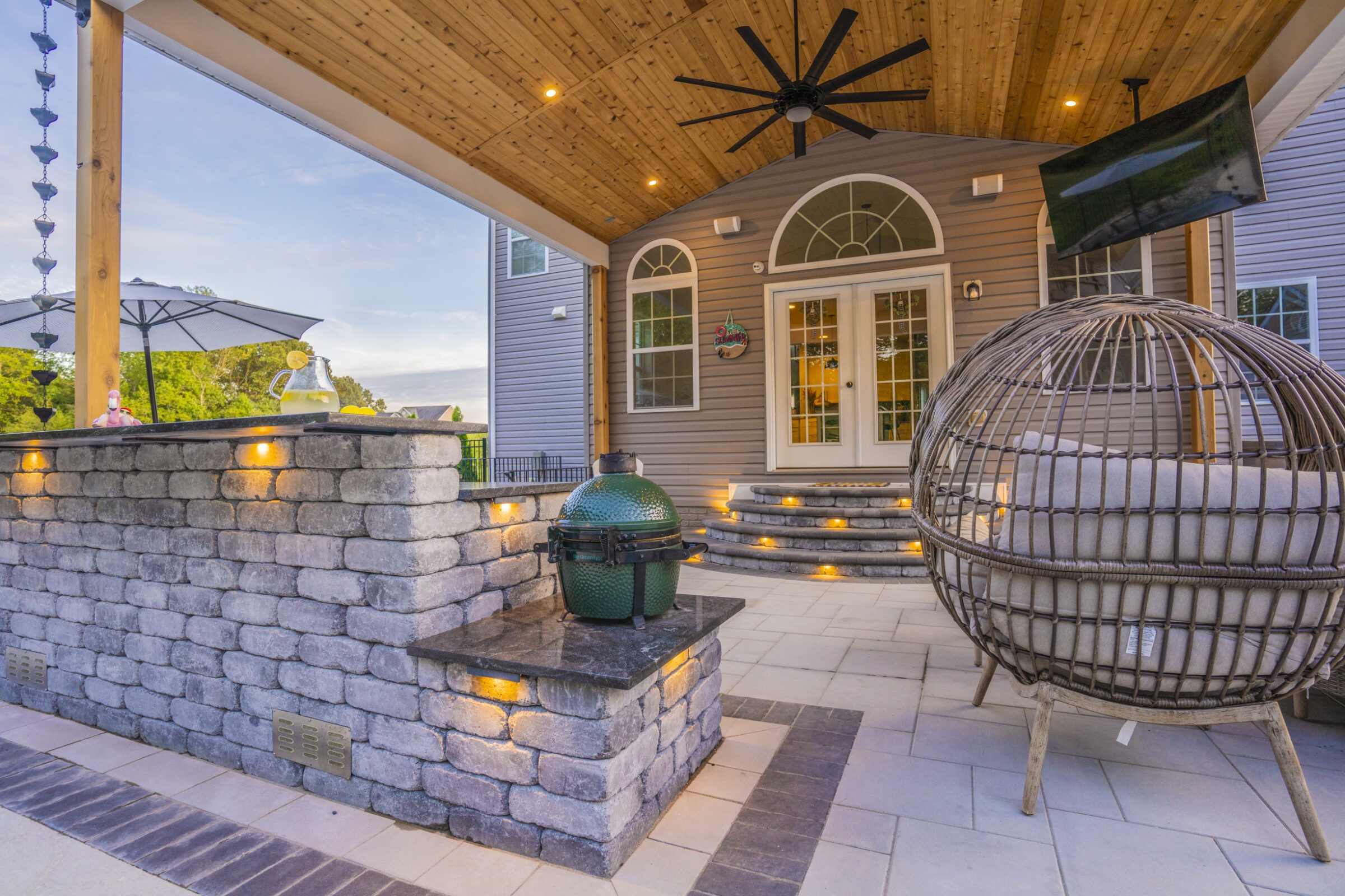Covered patio with stone grill, wicker seating, and decorative hanging lights. Glass doors lead inside. Outside, an umbrella table is visible.