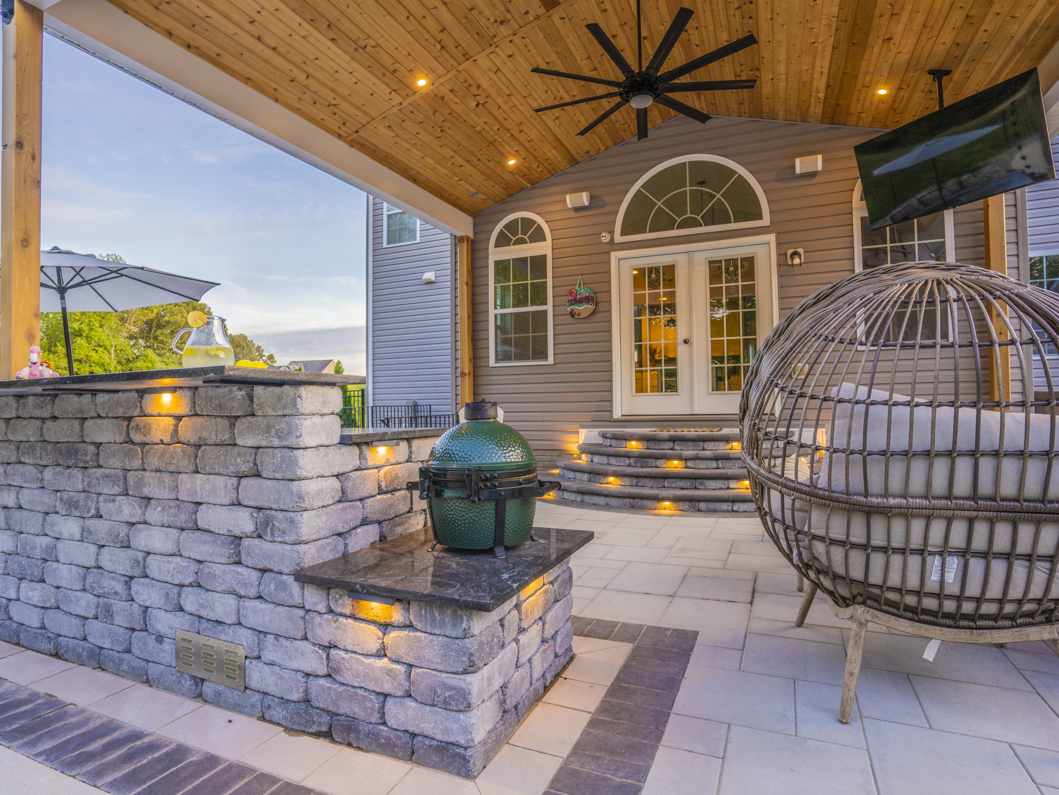 Covered patio with fan, brick wall, and grill. Egg chair and pitcher of lemonade create a cozy outdoor setting.