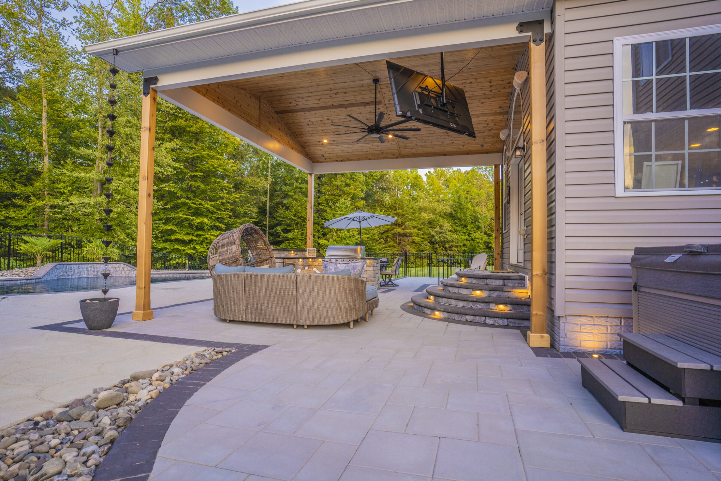 Modern patio with cozy seating, TV, fan, and fire pit. Surrounded by lush trees, poolside view, and glowing steps for elegant ambiance.