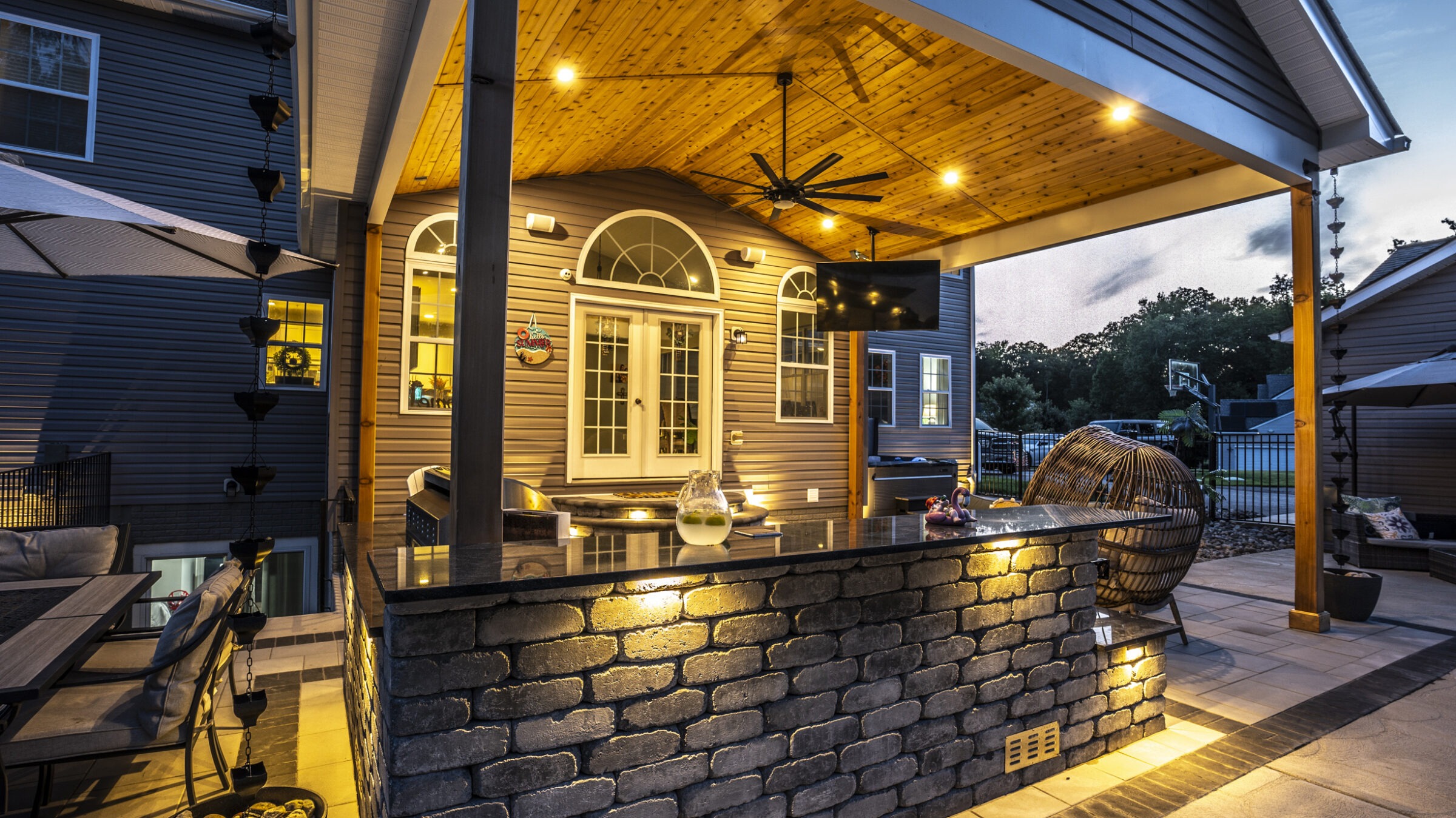 Modern outdoor patio featuring a stone bar, wicker chair, overhead lights, ceiling fan, and a television, adjacent to a house at dusk.