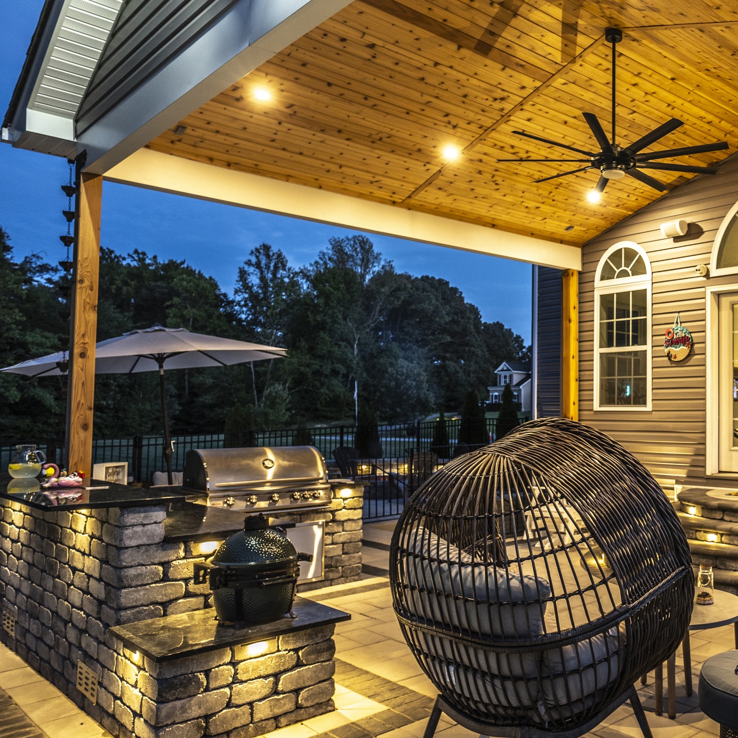 A cozy outdoor patio at dusk features seating, a grill, hanging lights, and surrounding trees, creating a warm, inviting atmosphere.