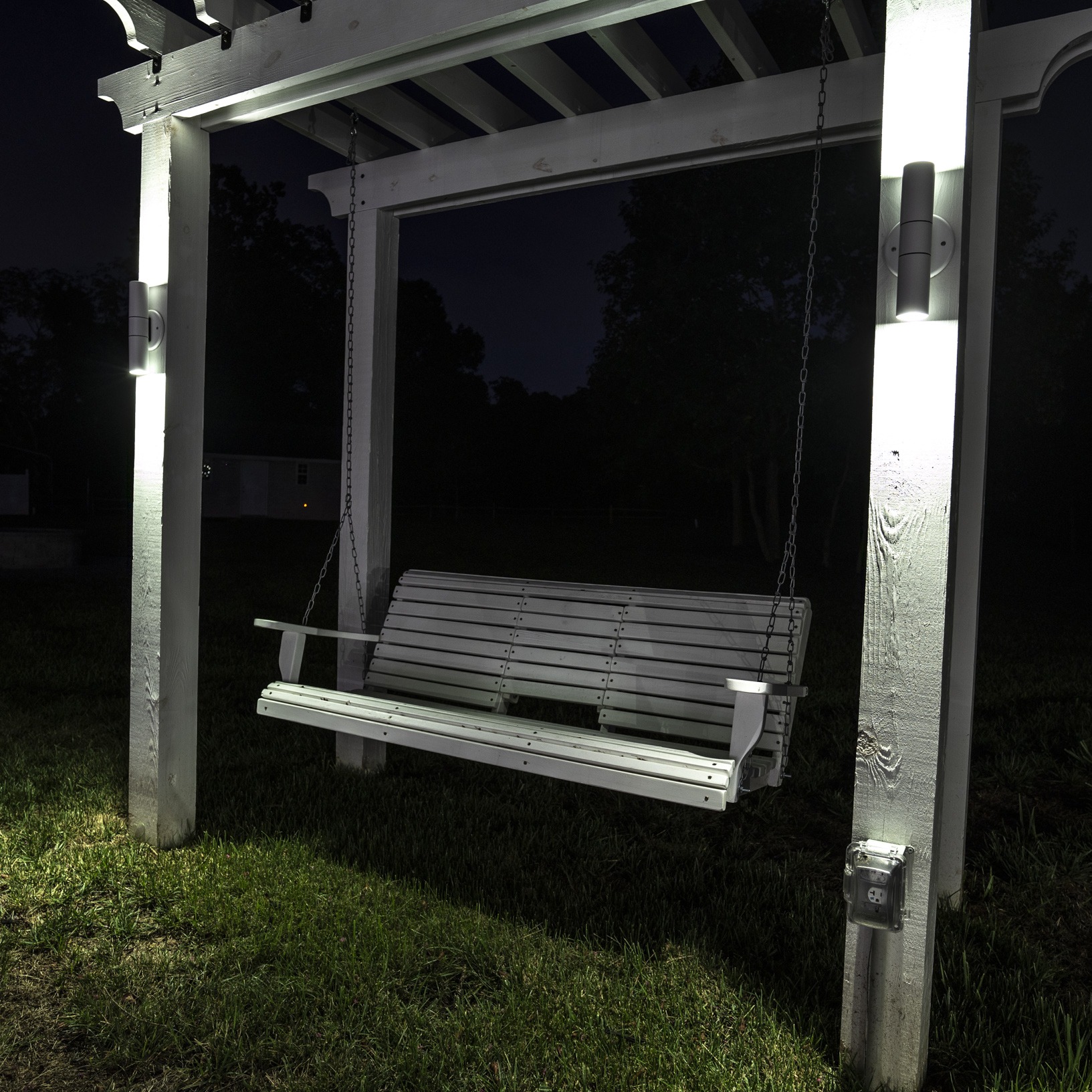A well-lit wooden swing hangs from a pergola at night, surrounded by grass and silhouetted trees in the background.