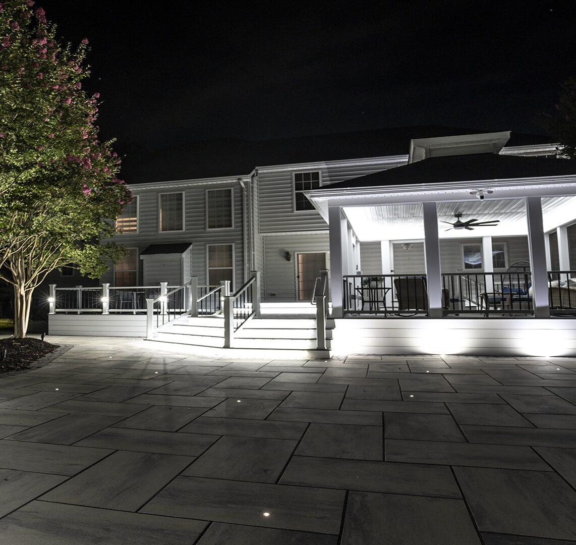 A well-lit house with a large outdoor patio and seating area at night, surrounded by trees under subtle garden lights.