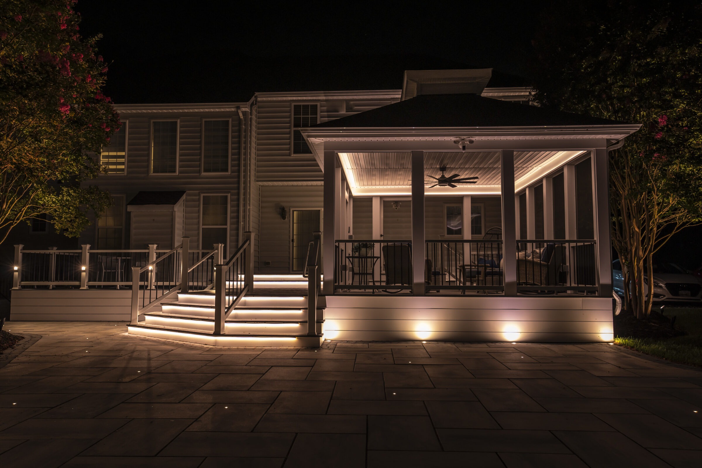 A house with illuminated steps and a covered patio at night, featuring a ceiling fan and surrounding trees, offers a warm ambiance.