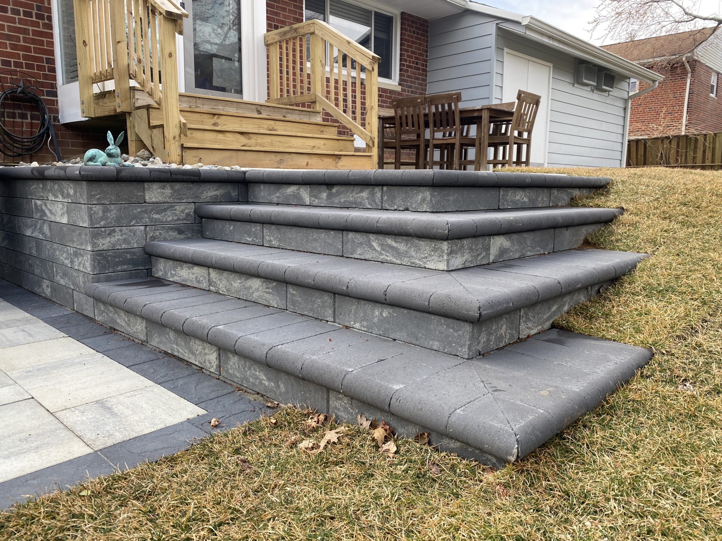 Gray stone steps lead to a wooden deck with outdoor dining furniture, adjacent to a brick house and grassy lawn.