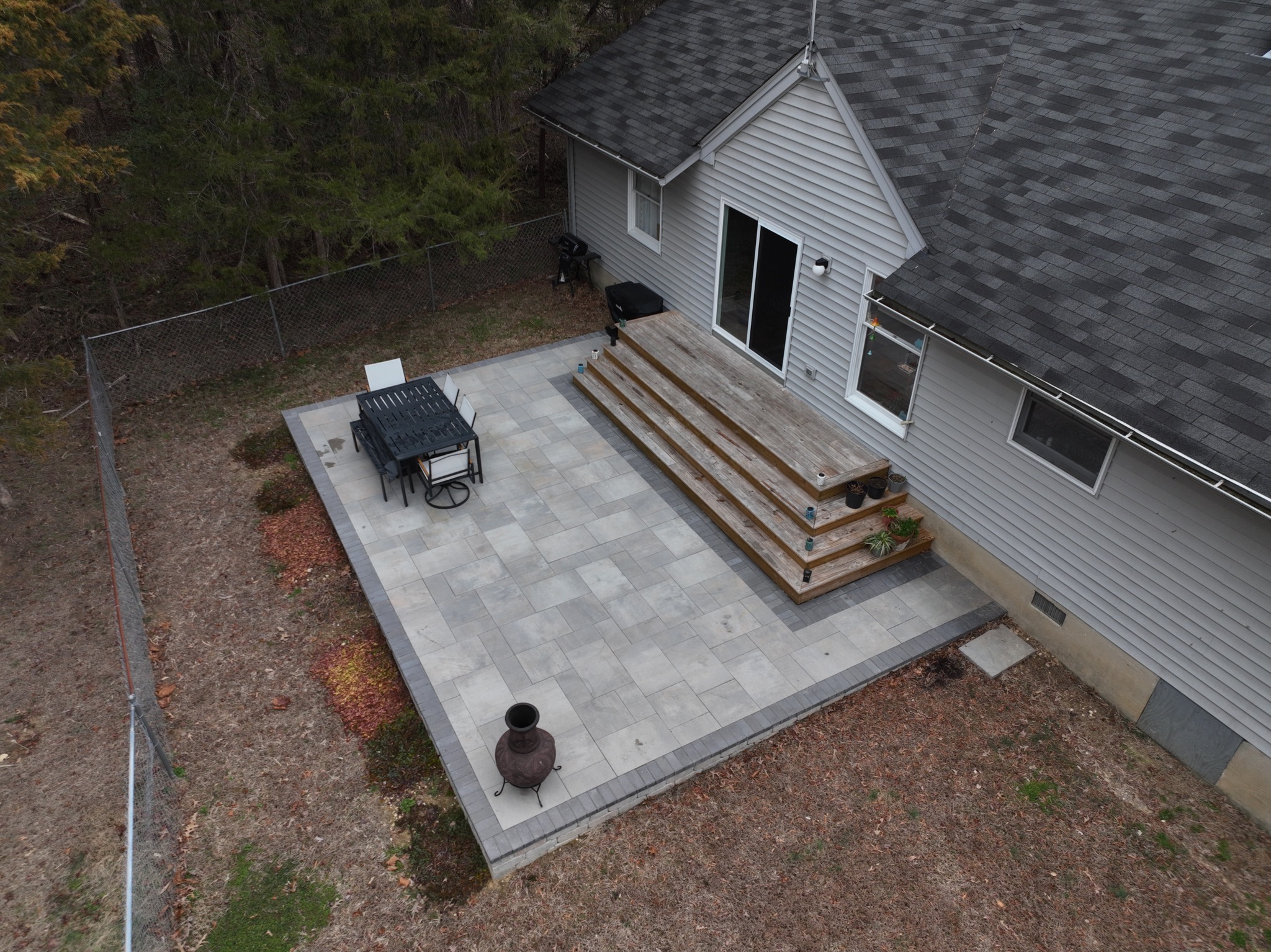 Aerial view of a house with a tiled patio, outdoor dining set, steps, and surrounding trees, creating a tranquil backyard space.