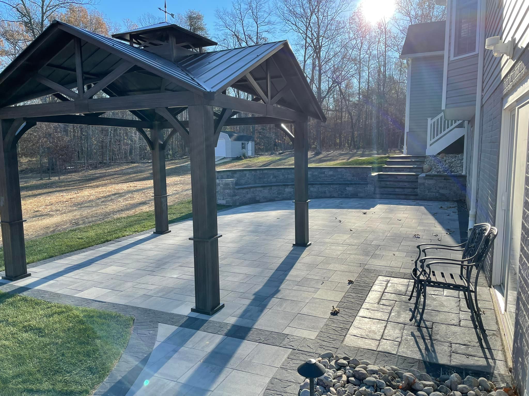 A backyard patio with pergola, stone pathway, and metal bench. Surrounded by woods under a bright sunlit sky, showing a residential house exterior.