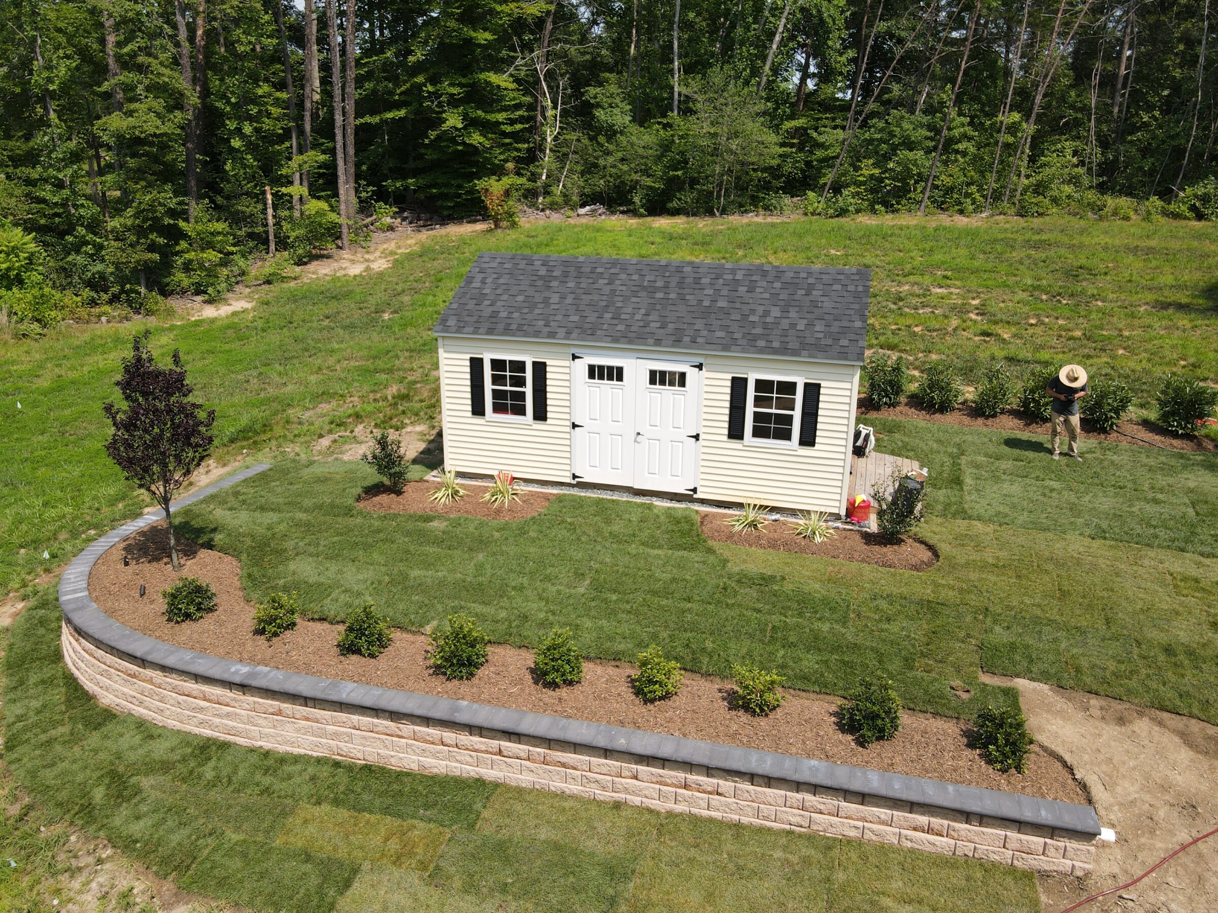 A small shed with two doors, surrounded by landscaped garden and trees. A person wearing a hat stands nearby.