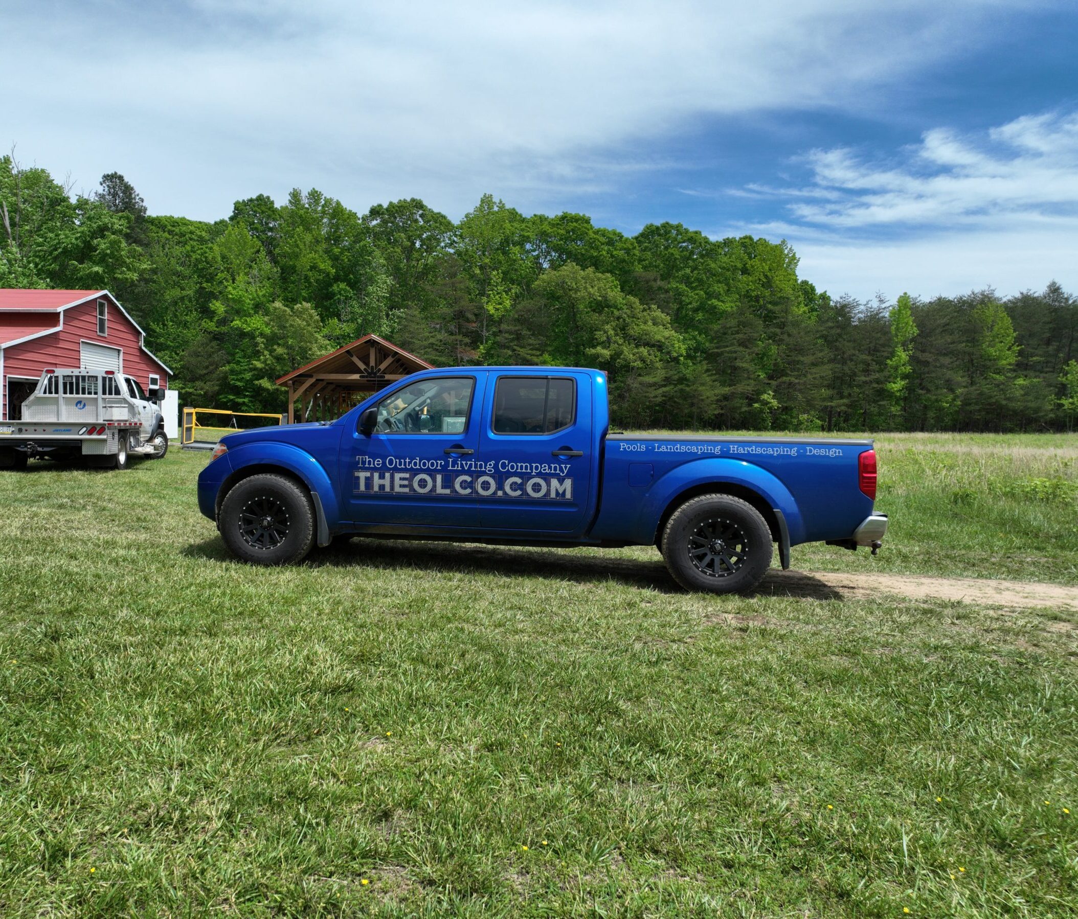 A blue pickup truck beside a red barn on a grassy field, surrounded by trees under a partly cloudy sky.