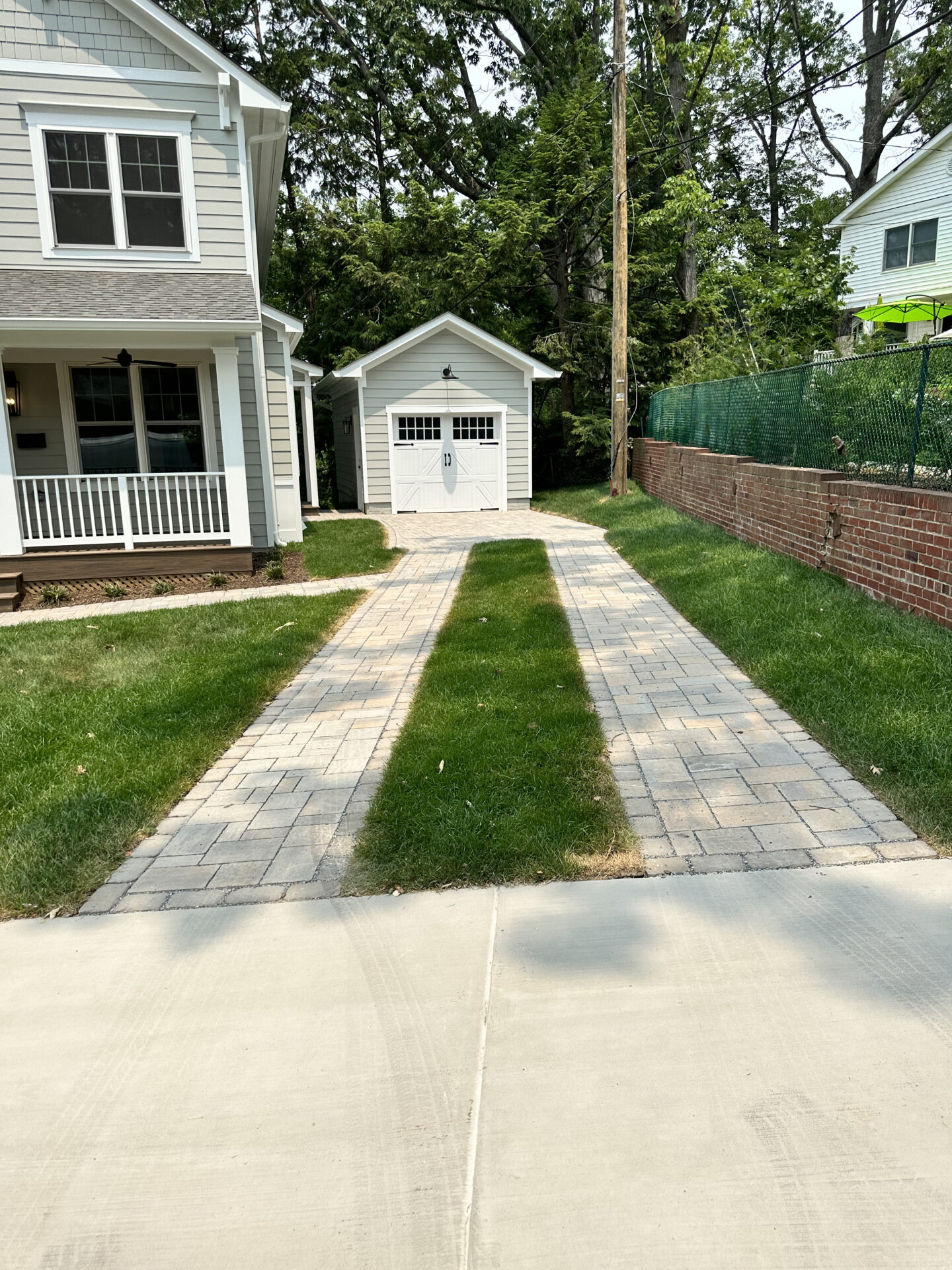A suburban driveway leads to a small garage, flanked by a gray house and a brick wall with surrounding trees.