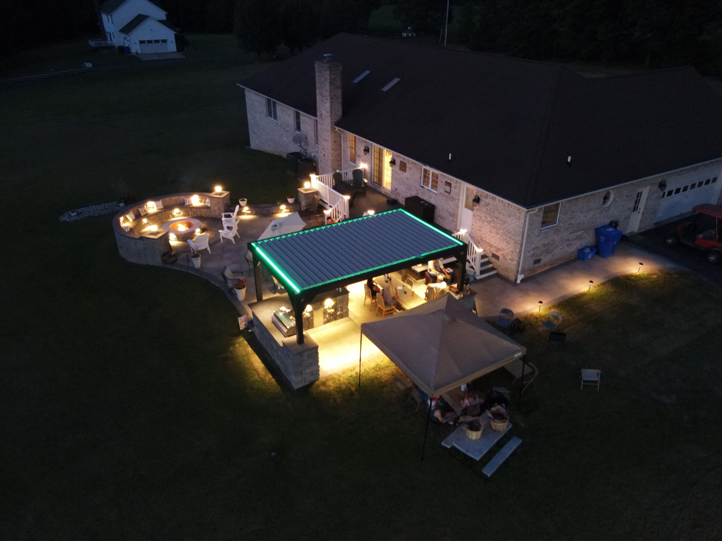 Aerial view of a house with a well-lit patio area, pergola, and seating. Nighttime setting with surrounding lawn and trees.
