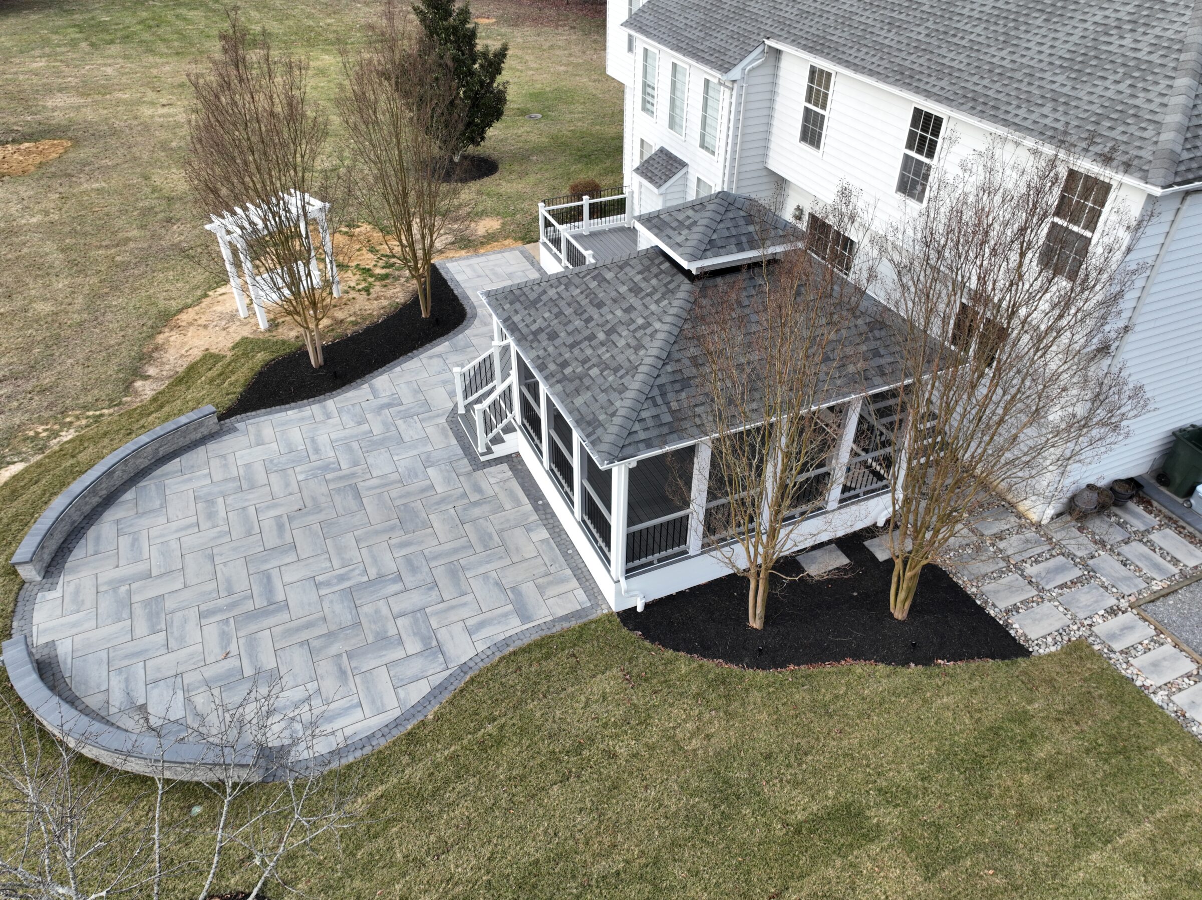 Aerial view of a house with a large paved patio, white pergola, and landscaped garden with bare trees on grass.