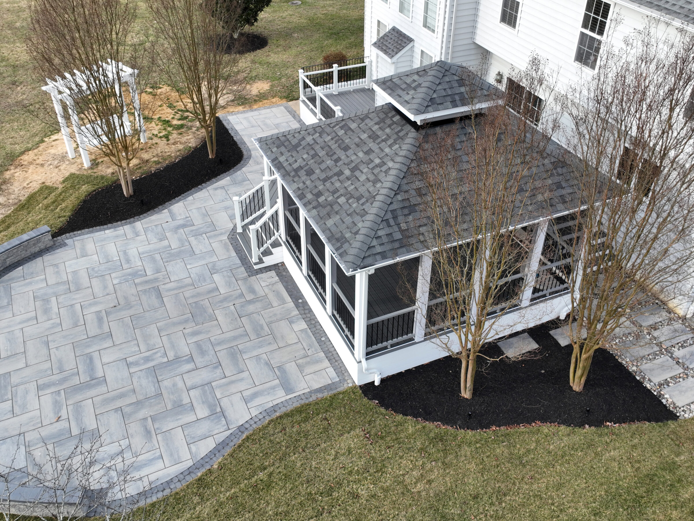 Aerial view of a modern backyard featuring a paved patio, landscaped garden, white arbor, and trees beside a residential house.