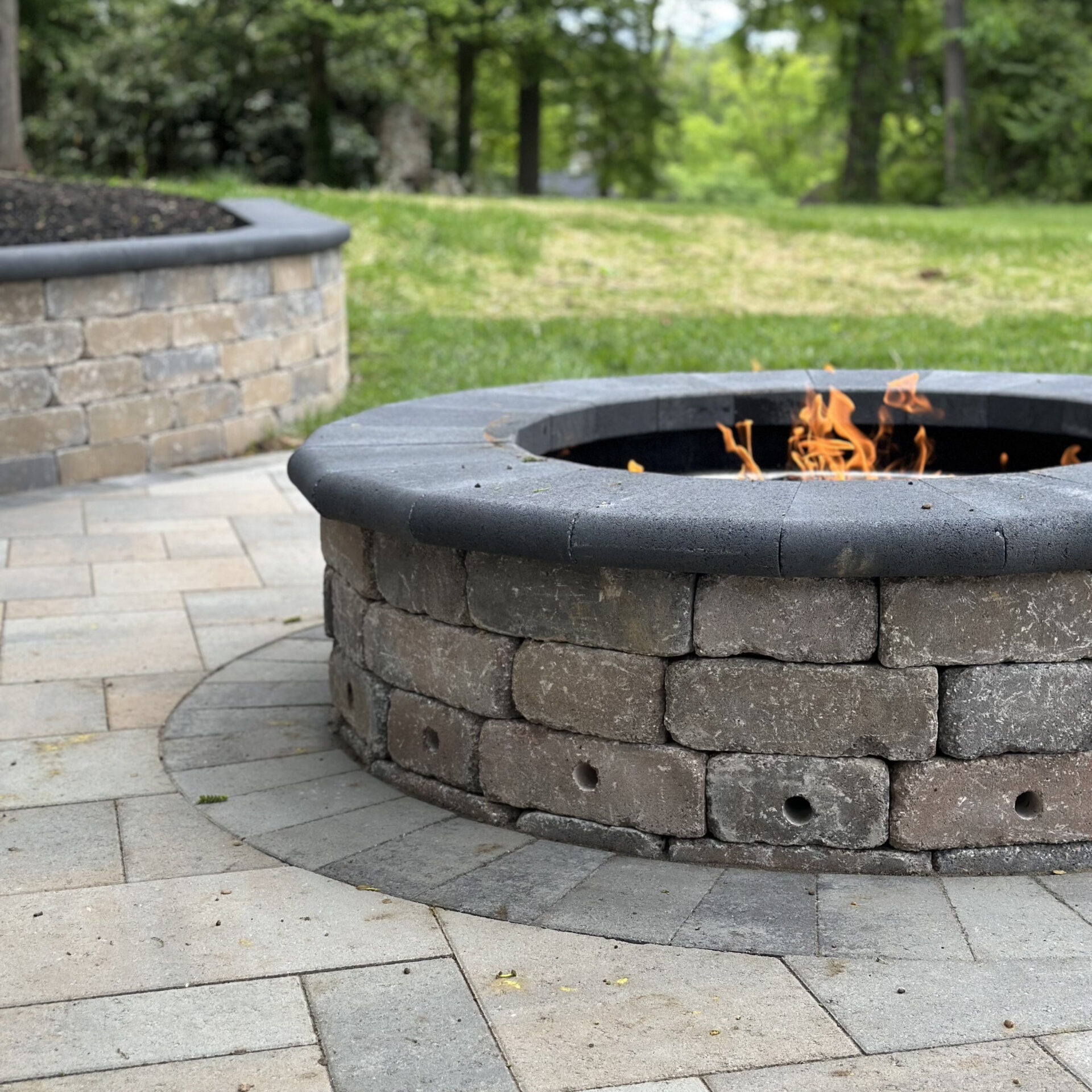 A stone fire pit with flames surrounded by a paved patio and grassy area, set in a wooded outdoor setting.