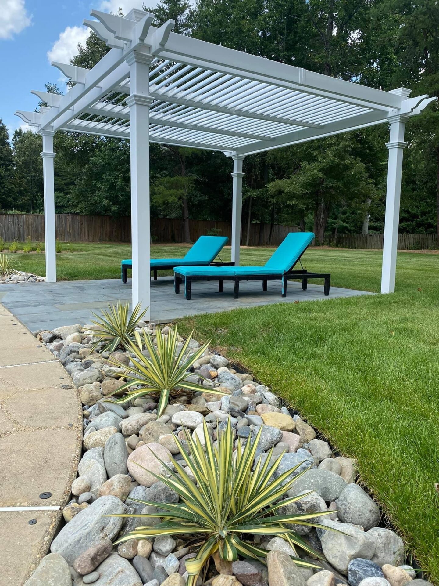 A white pergola with blue lounge chairs on a lawn. Nearby, decorative stones and plants line a walkway, surrounded by trees and wooden fence.
