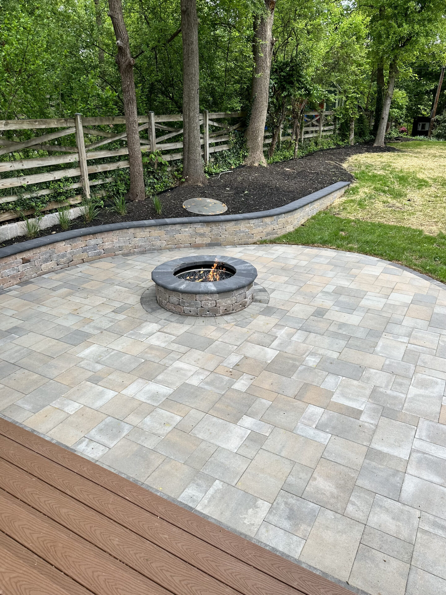 A backyard patio with a circular stone fire pit, surrounded by trees and wooden fence, creates a peaceful outdoor setting.