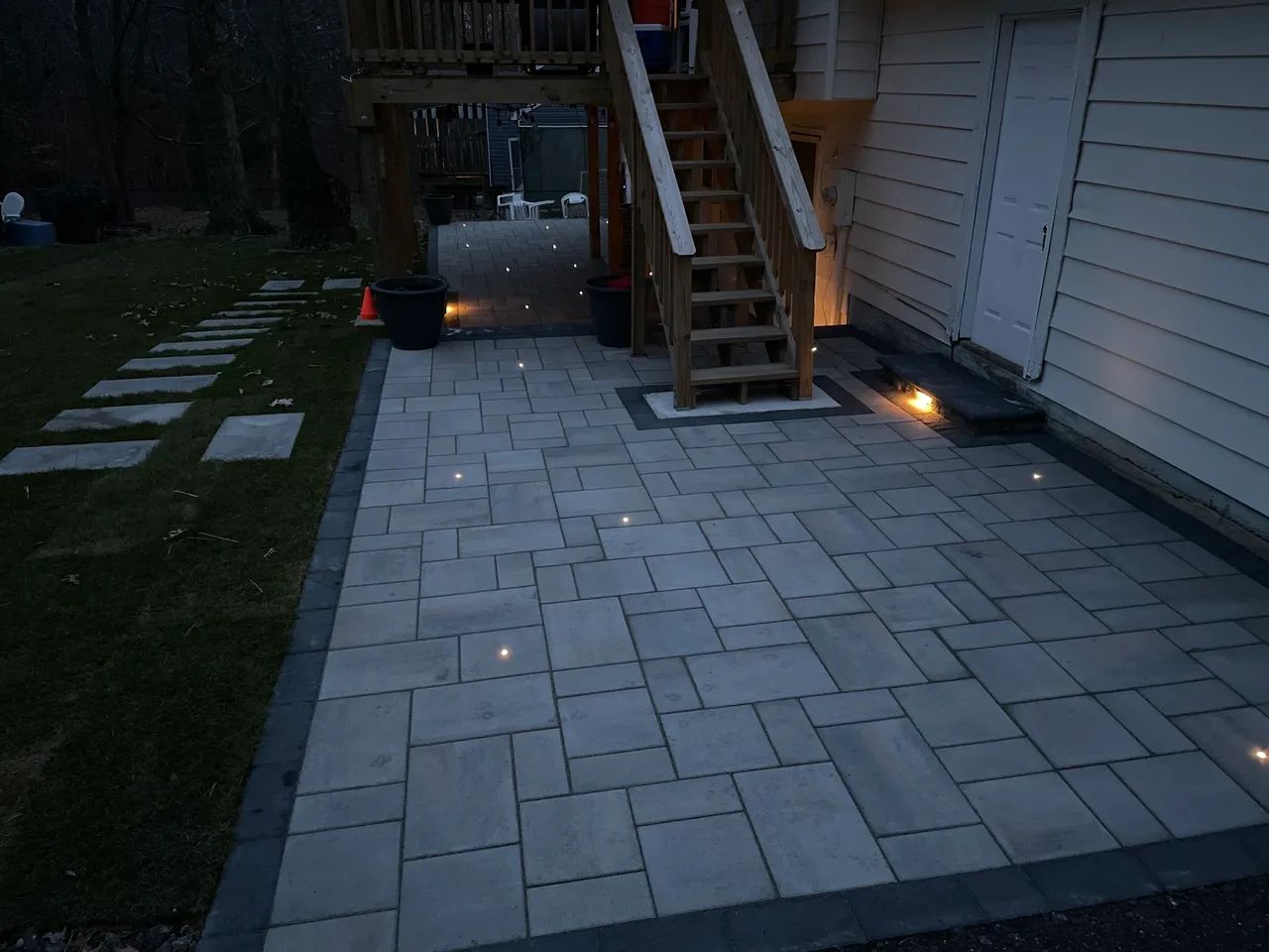 A patio with stone tiles and embedded lights, adjacent to a wooden staircase, illuminated softly in an evening setting.