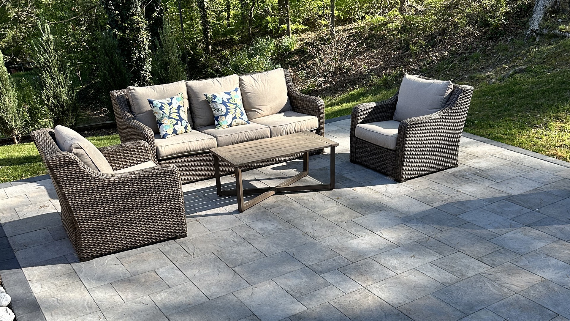 Outdoor stone patio with wicker furniture set, including a sofa, armchairs, and a wooden table, surrounded by lush greenery.