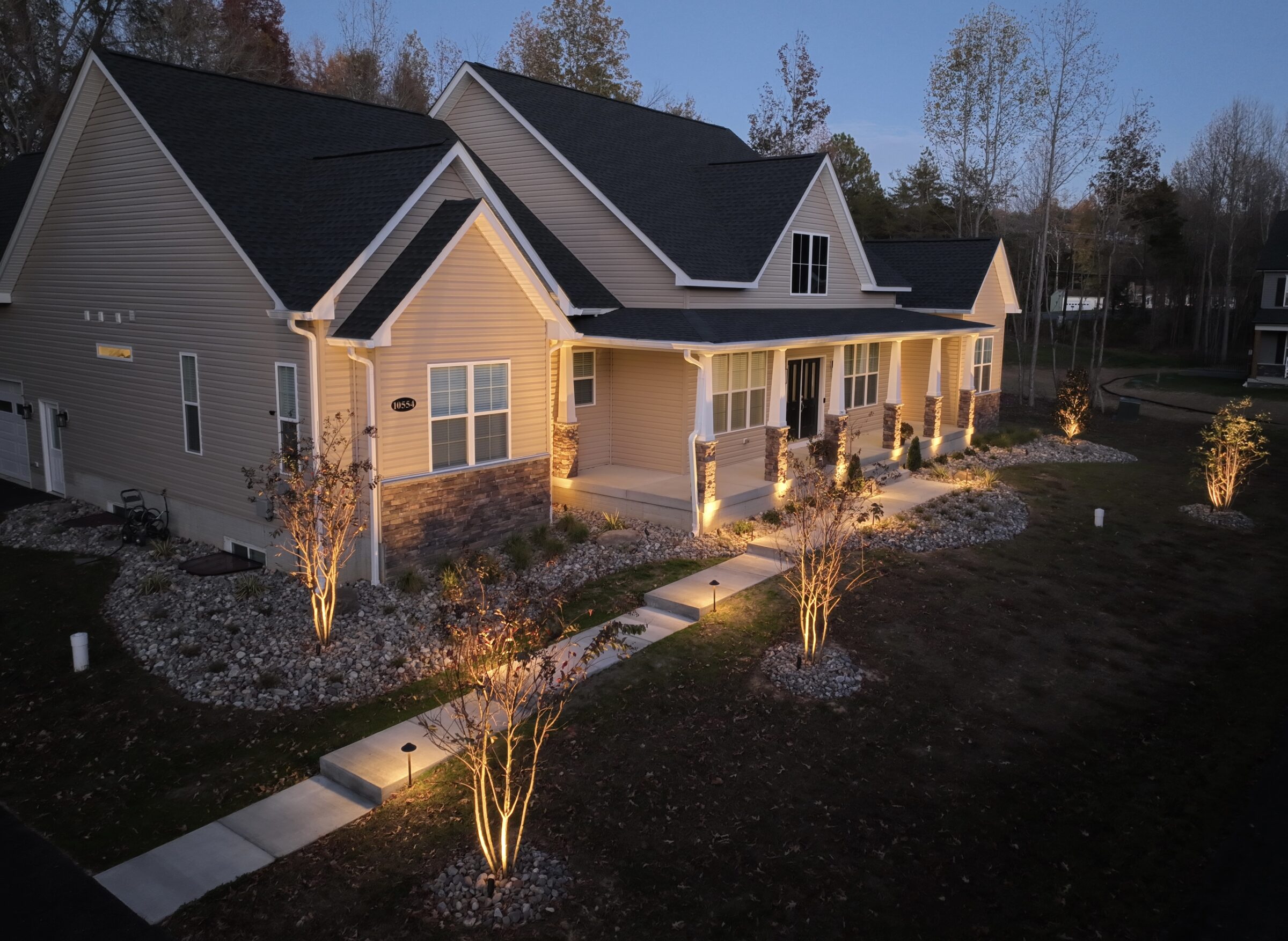 Single-story house with illuminated pathways, surrounded by trees and landscaped gardens, under a twilight sky. Cozy, inviting ambiance in a suburban setting.