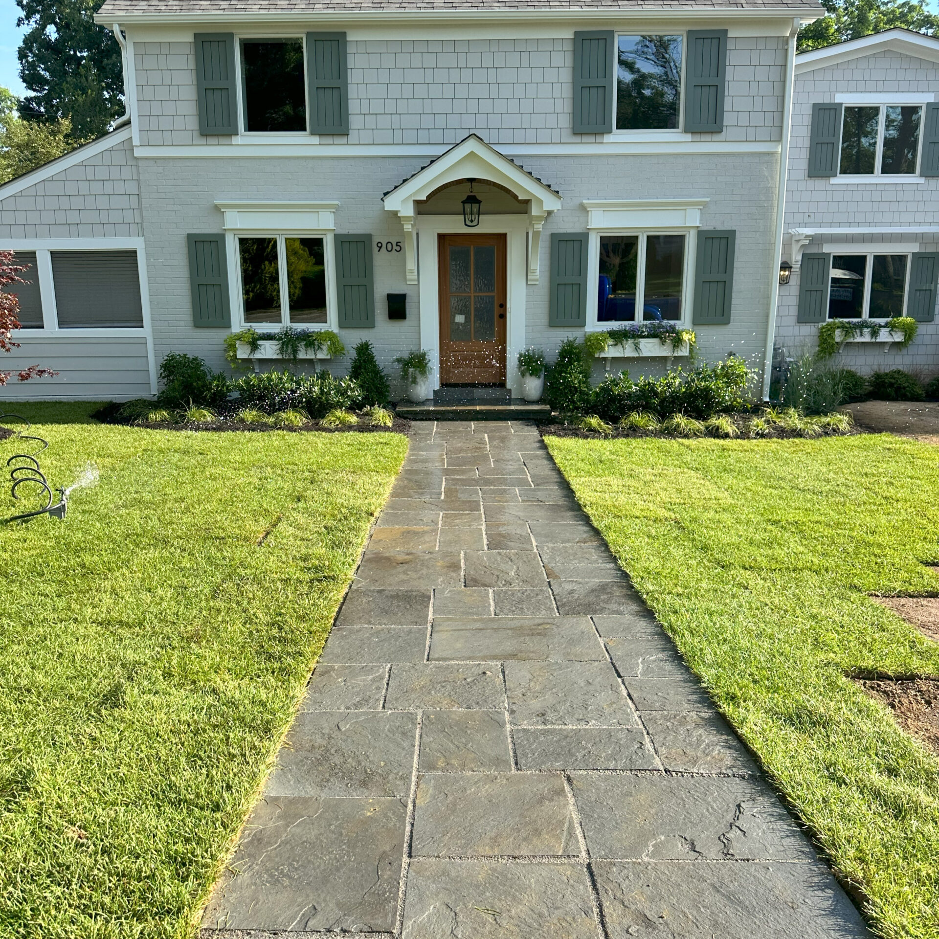 A two-story suburban house with gray siding, green shutters, and a tidy pathway through a well-maintained lawn, beneath a sunny sky.